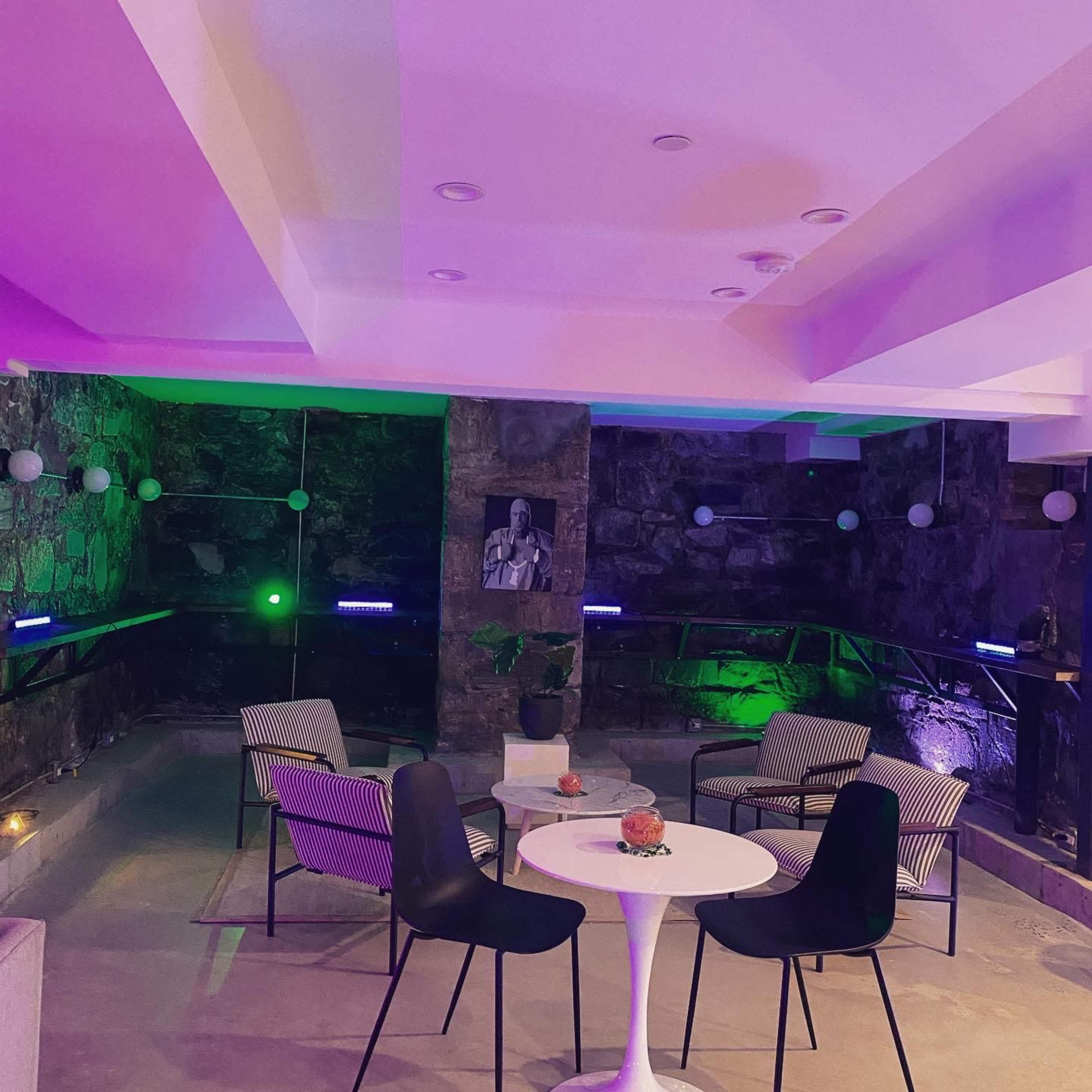 A modern lounge area features a round table surrounded by four chairs, set against a backdrop of stone walls illuminated in green and purple light.