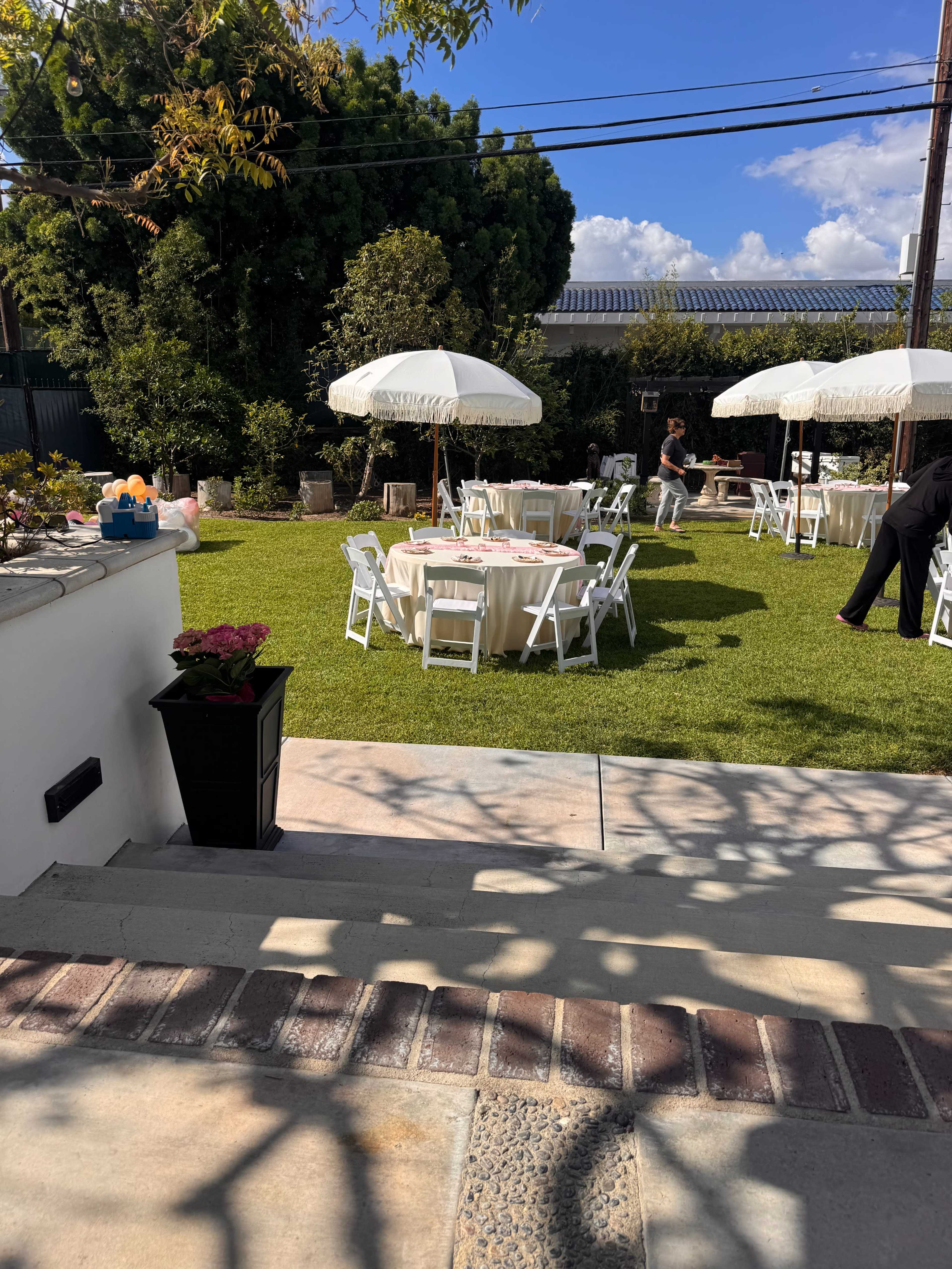A well-set outdoor space features several round tables with white tablecloths and umbrellas on a green lawn, with an individual preparing for an event in the background.