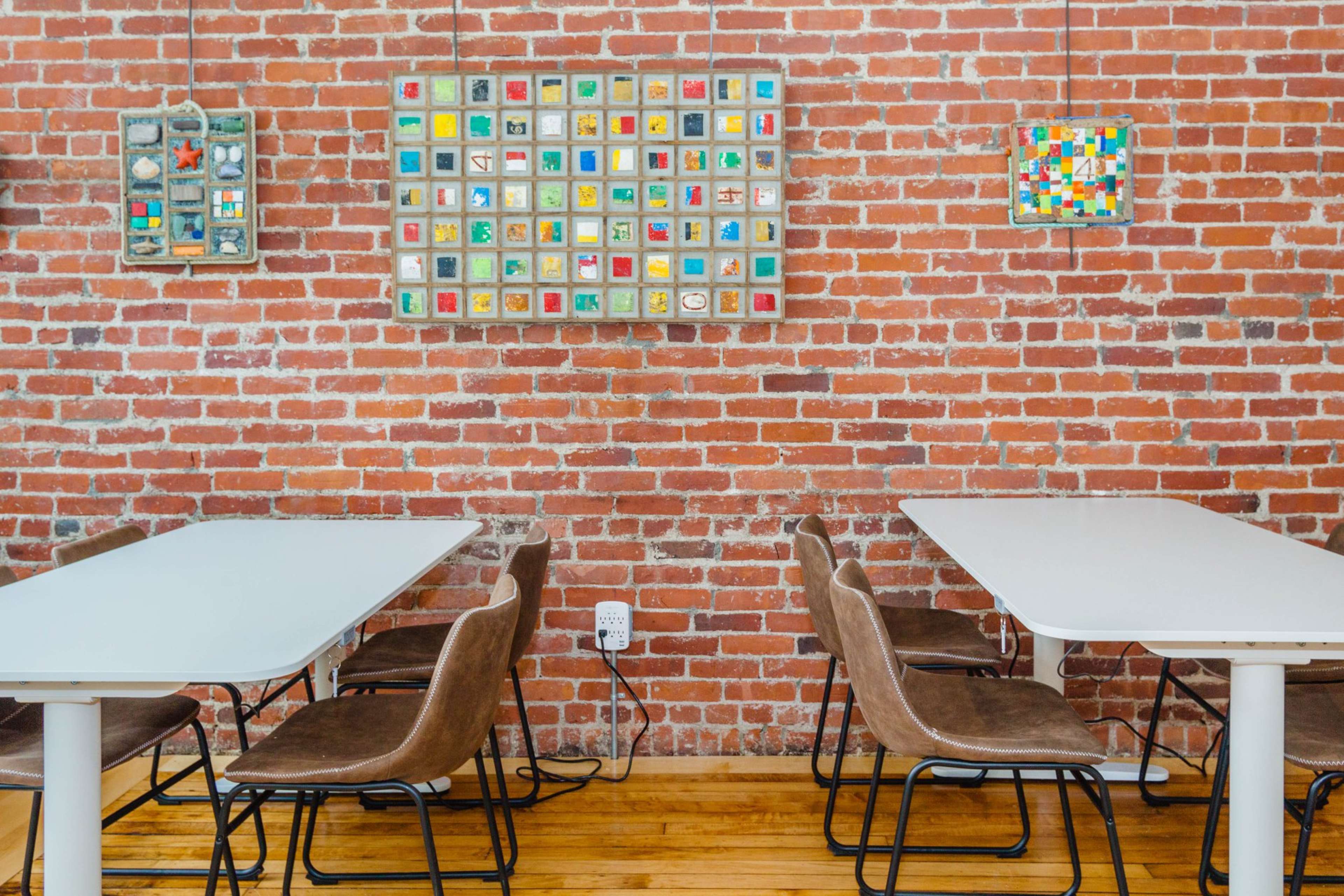 A modern café interior featuring white tables and brown chairs against a brick wall adorned with colorful tile artwork.