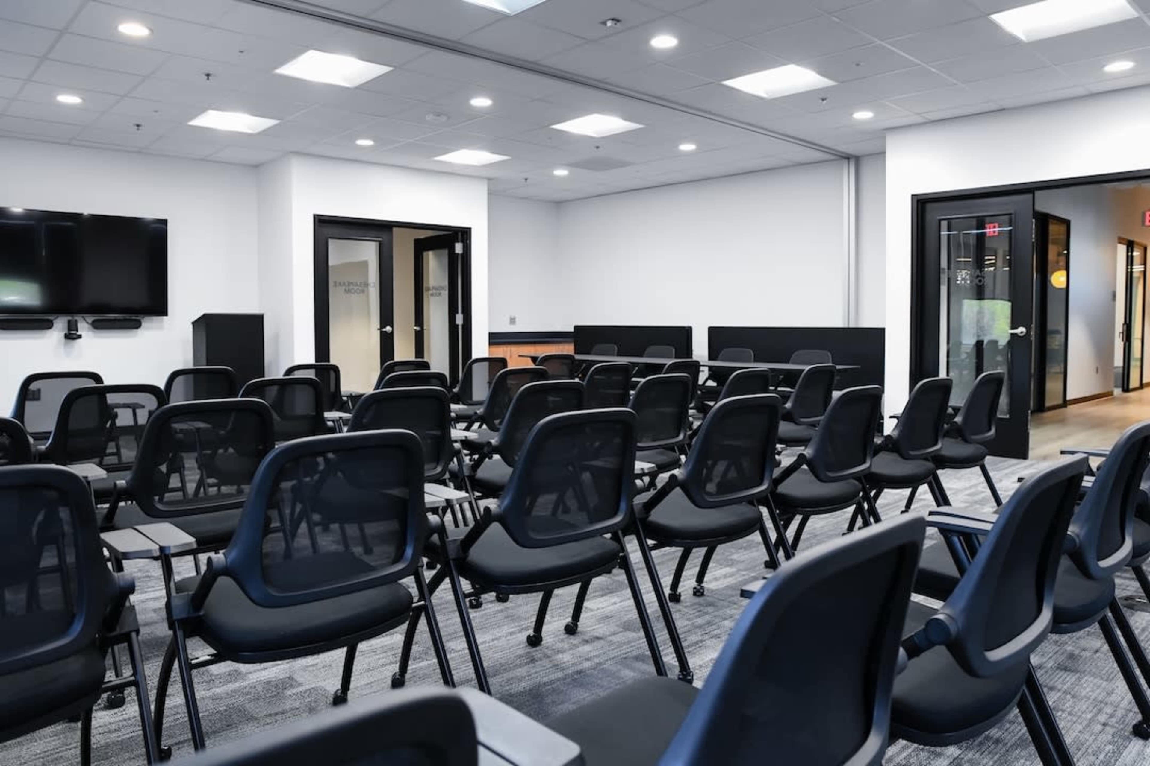 The image shows a modern conference room set up with rows of black chairs facing a presentation area and large screen.