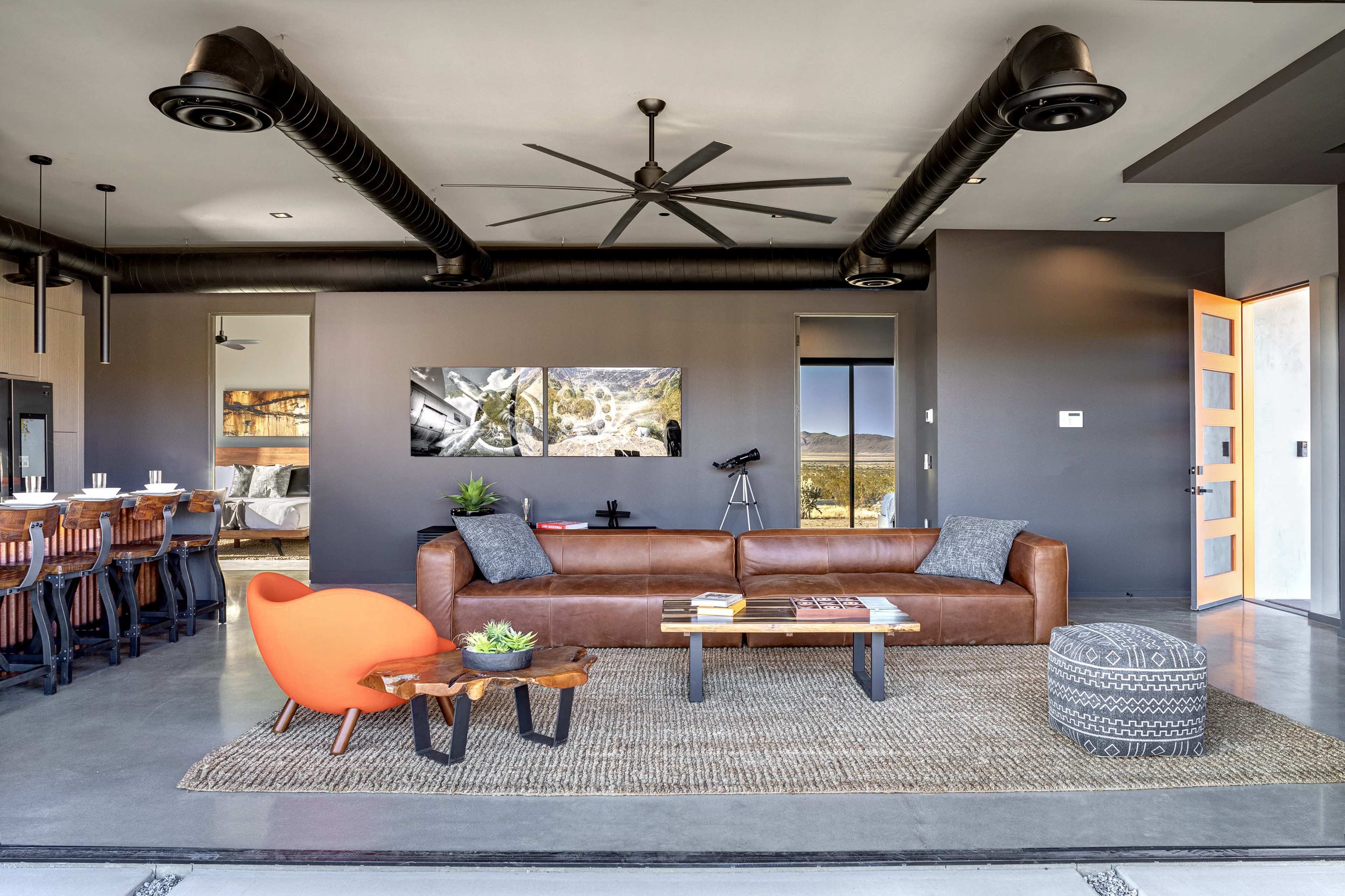 The image shows a modern living room featuring a brown leather sofa, an orange accent chair, a wooden coffee table, and a large doorway leading outdoors, with exposed ventilation pipes and a dining area in the background.
