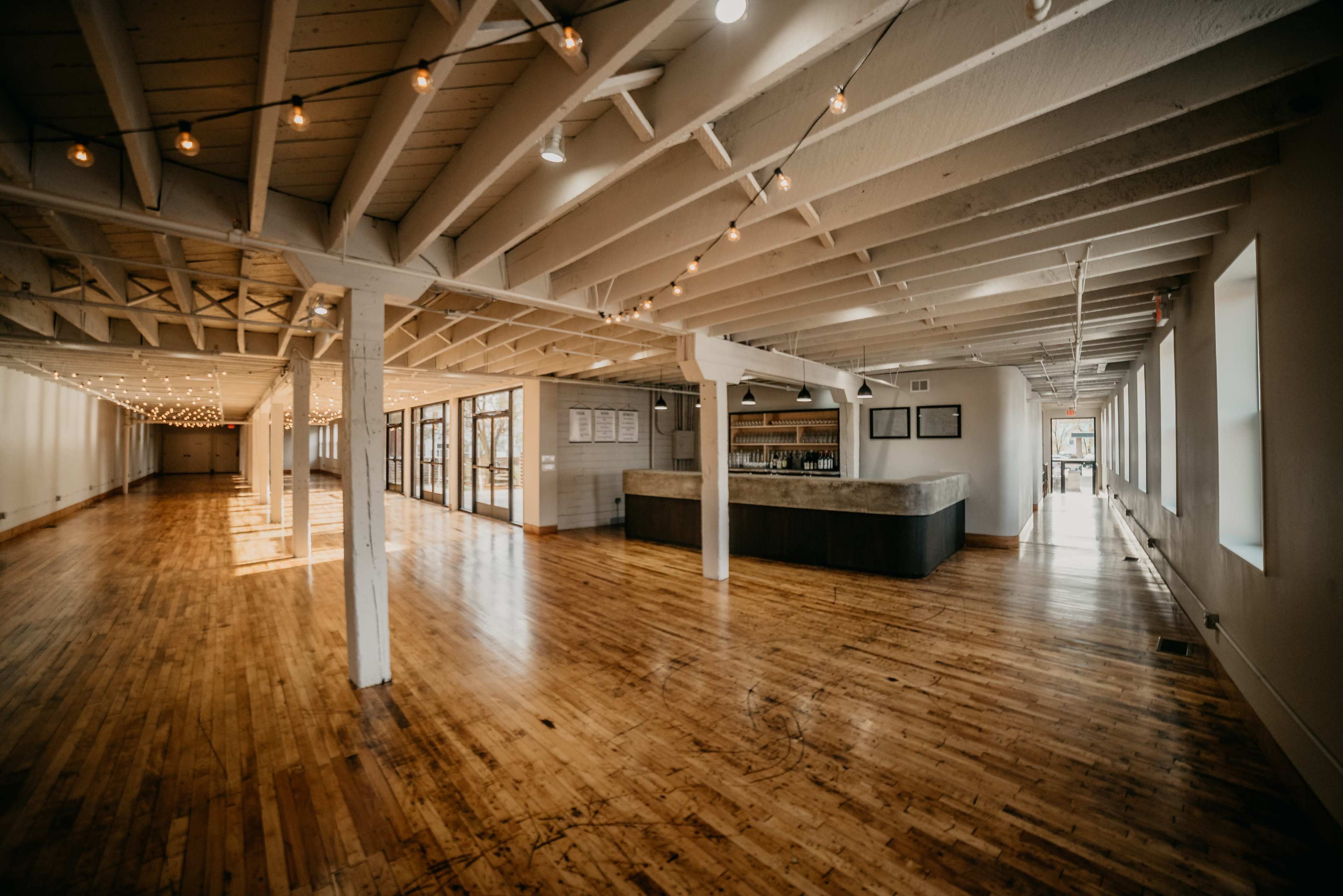 The image shows a spacious, empty interior with wooden floors and exposed beams, featuring a long counter at one end and string lights overhead.