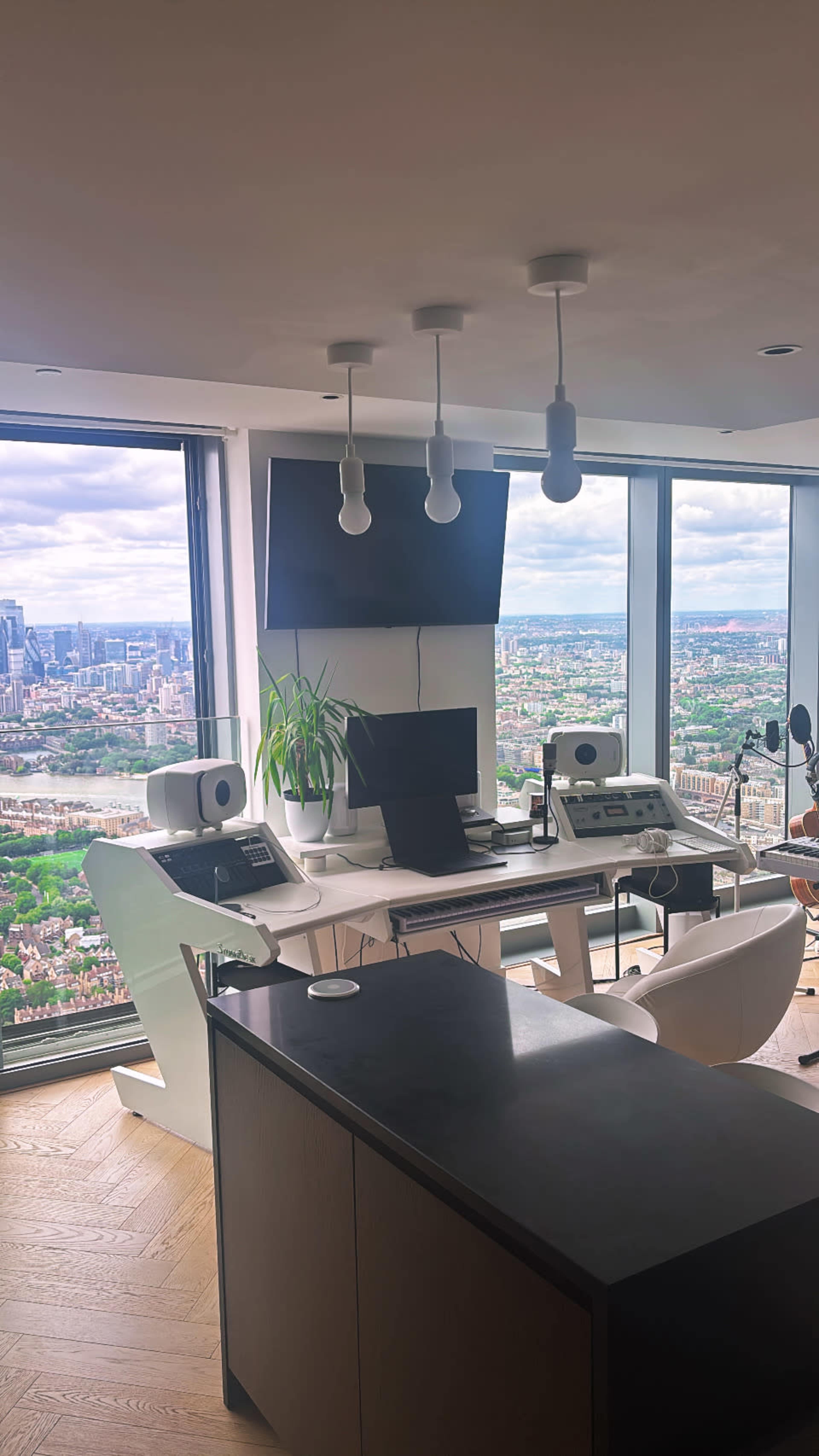 A modern workspace featuring a sleek desk with dual monitors, audio equipment, and a large window that offers a panoramic view of the city skyline.