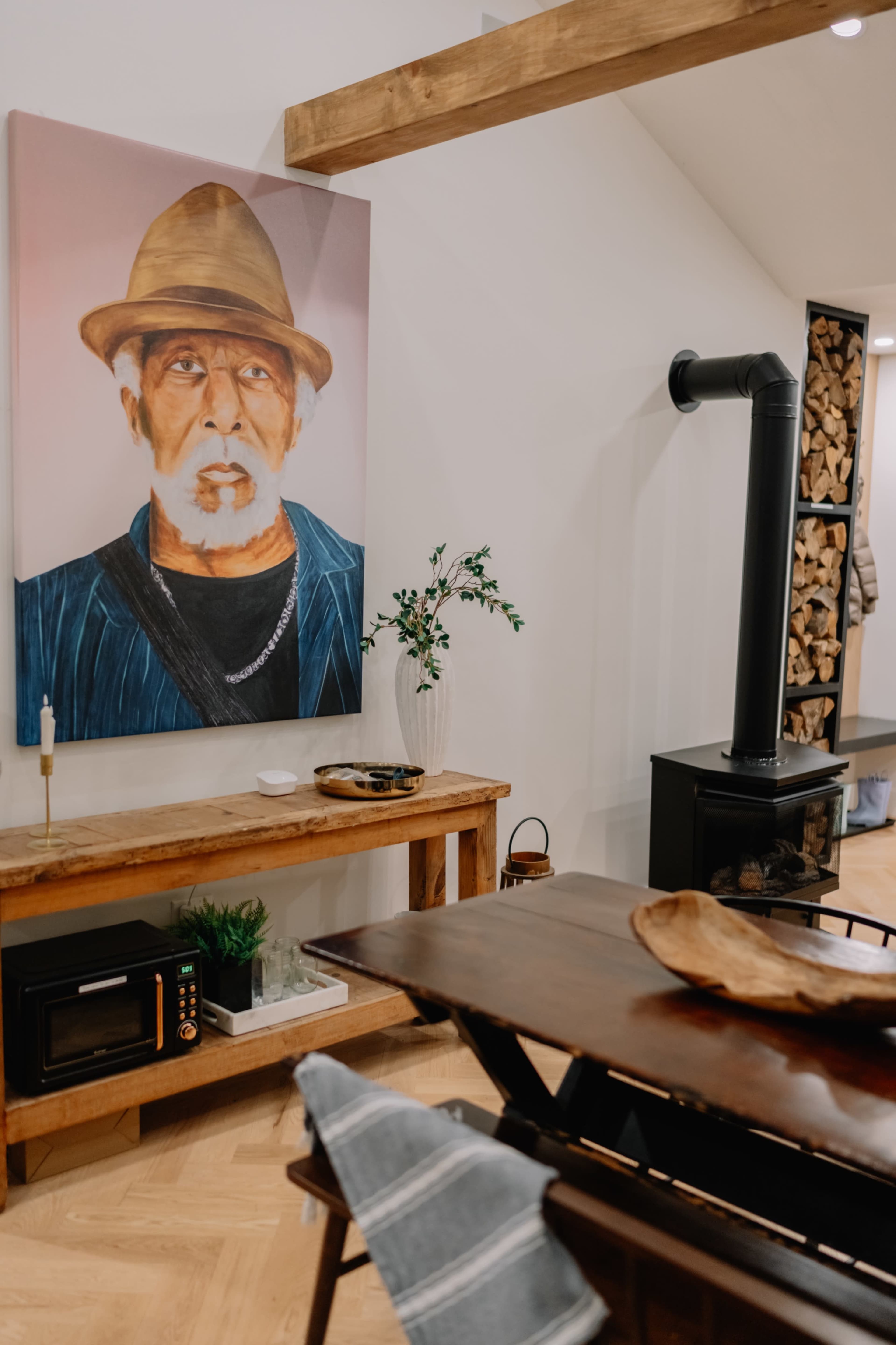The image shows a cozy interior with a wooden table, a portrait of a man in a hat on the wall, a black stove, and neatly stacked firewood in a corner.