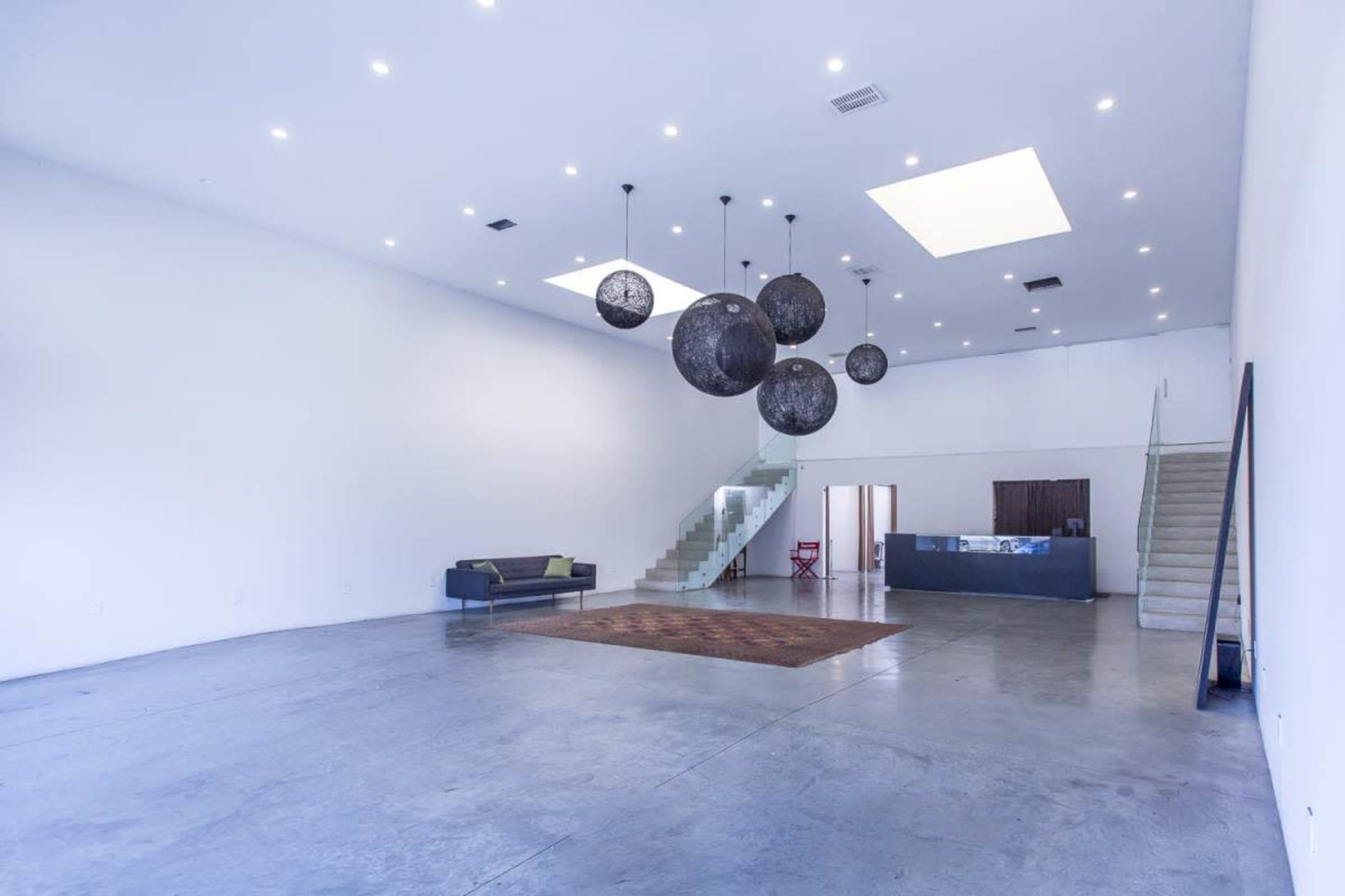 The image shows a spacious, minimalist room with high ceilings, a sofa, a couple of large decorative light fixtures, a reception area, and a staircase.