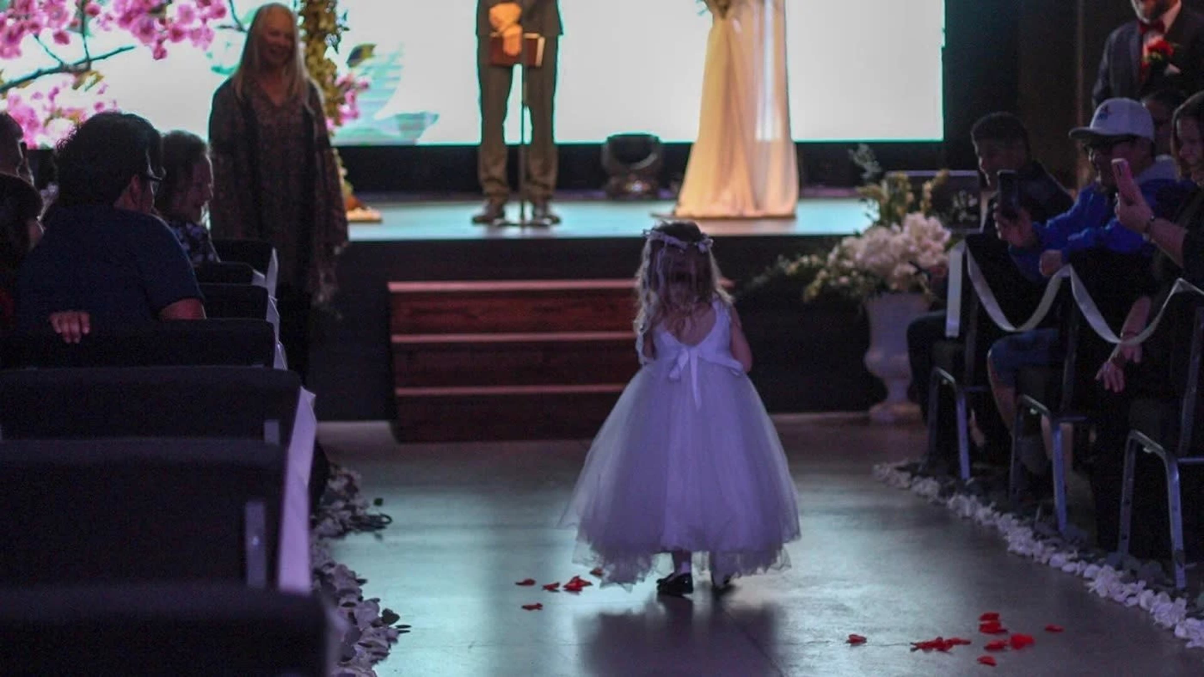 A young girl in a white dress walks down an aisle lined with chairs and flower petals toward a couple standing at the front of a wedding venue.