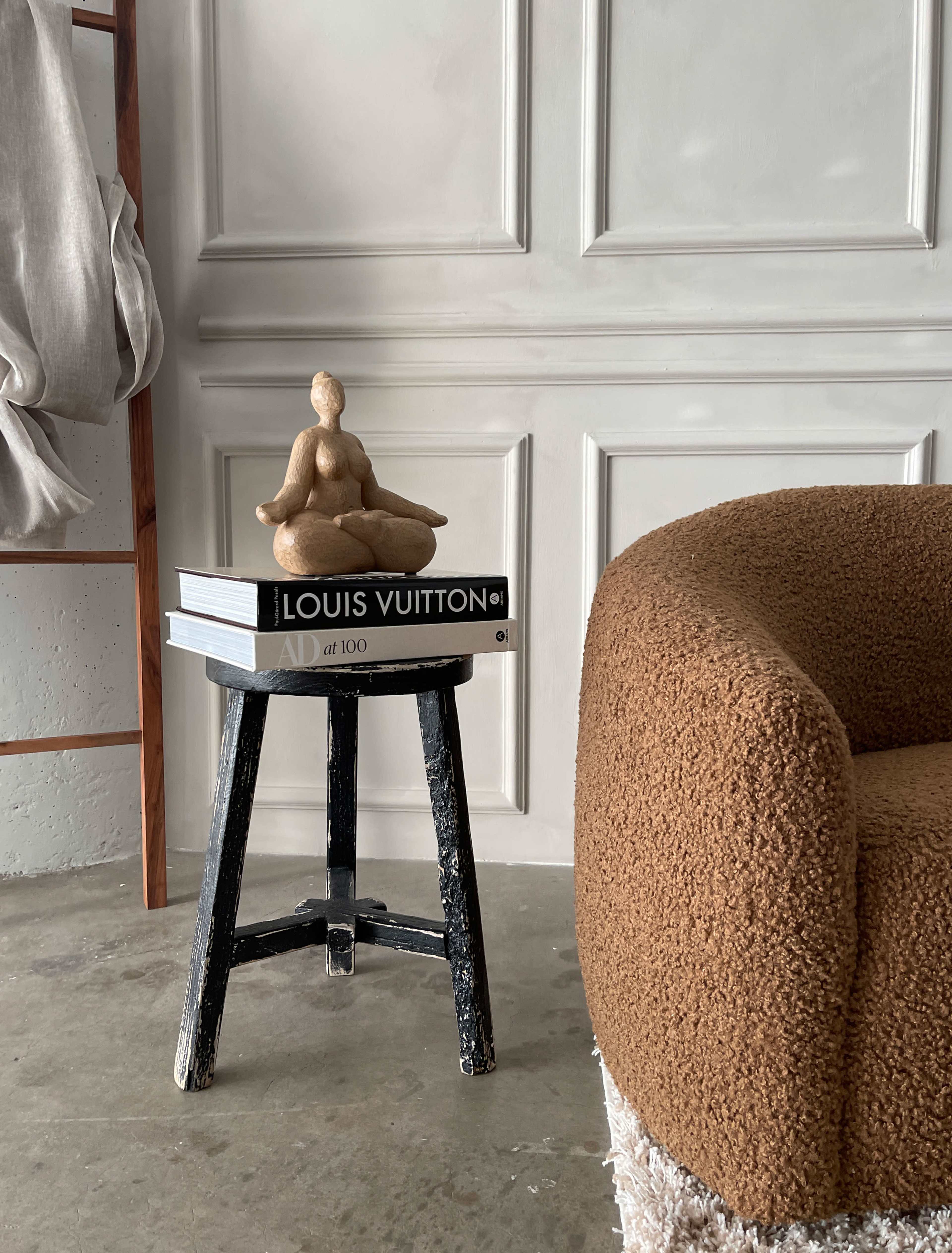 A textured brown chair sits next to a black stool stacked with books and topped by a sculpture against a white paneled wall.