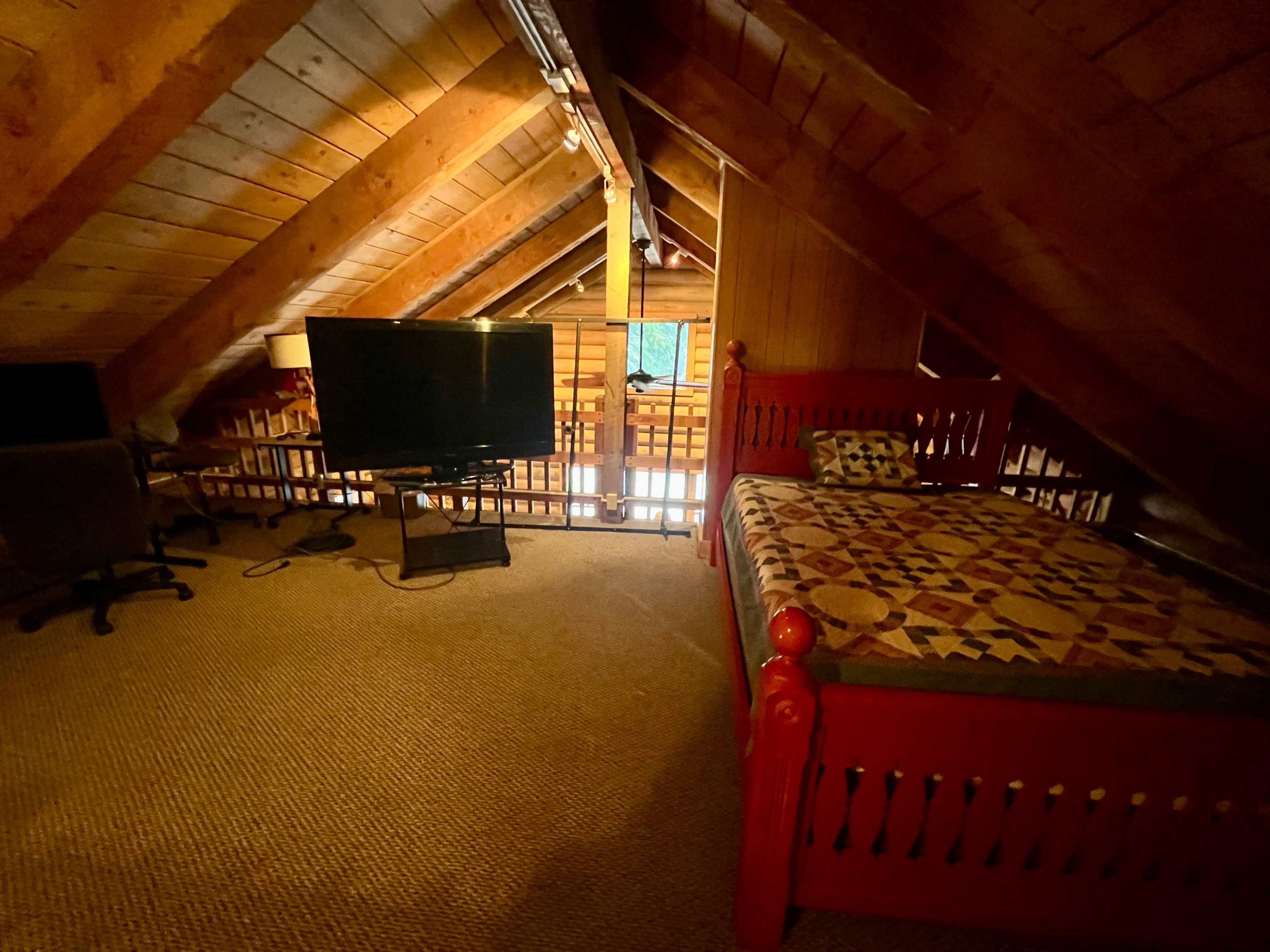 An attic bedroom featuring a wooden bed with a quilt, a large TV on a stand, and a carpeted floor.
