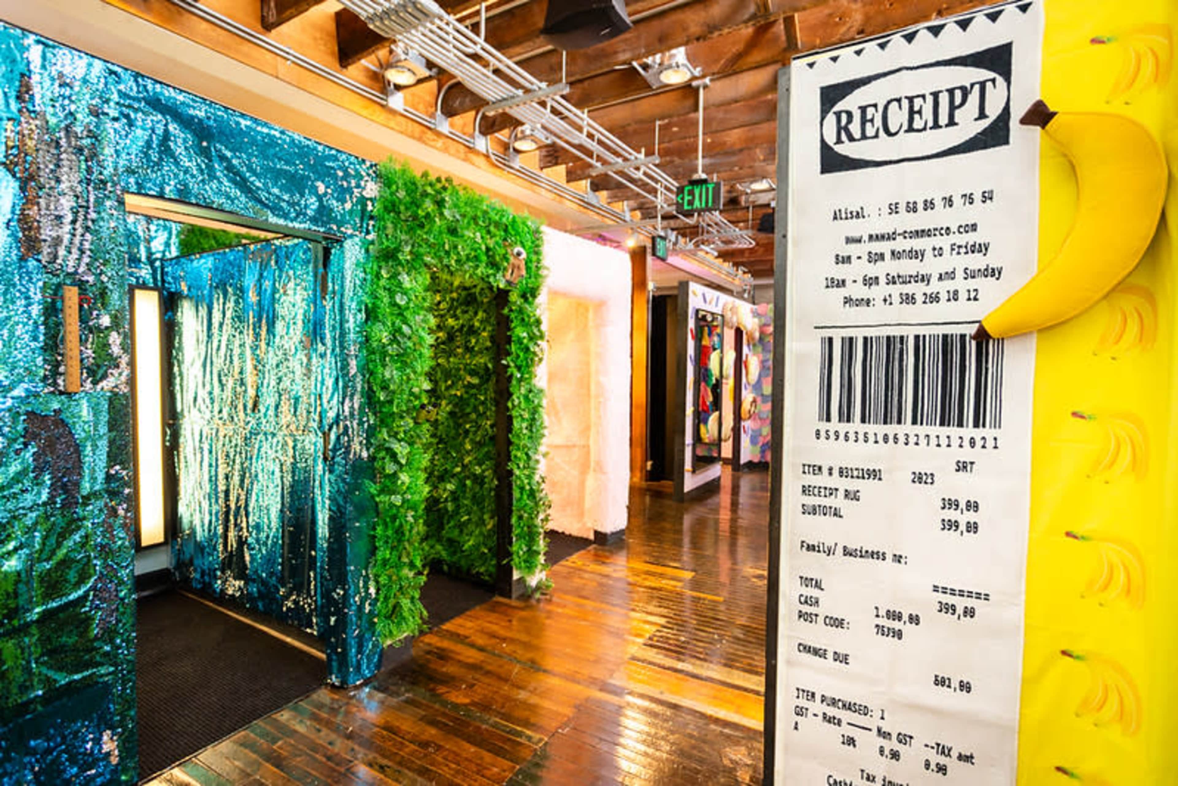 The image shows a vibrant indoor space with green foliage decor, a shimmering wall, and colorful artworks displayed along the wooden floor.