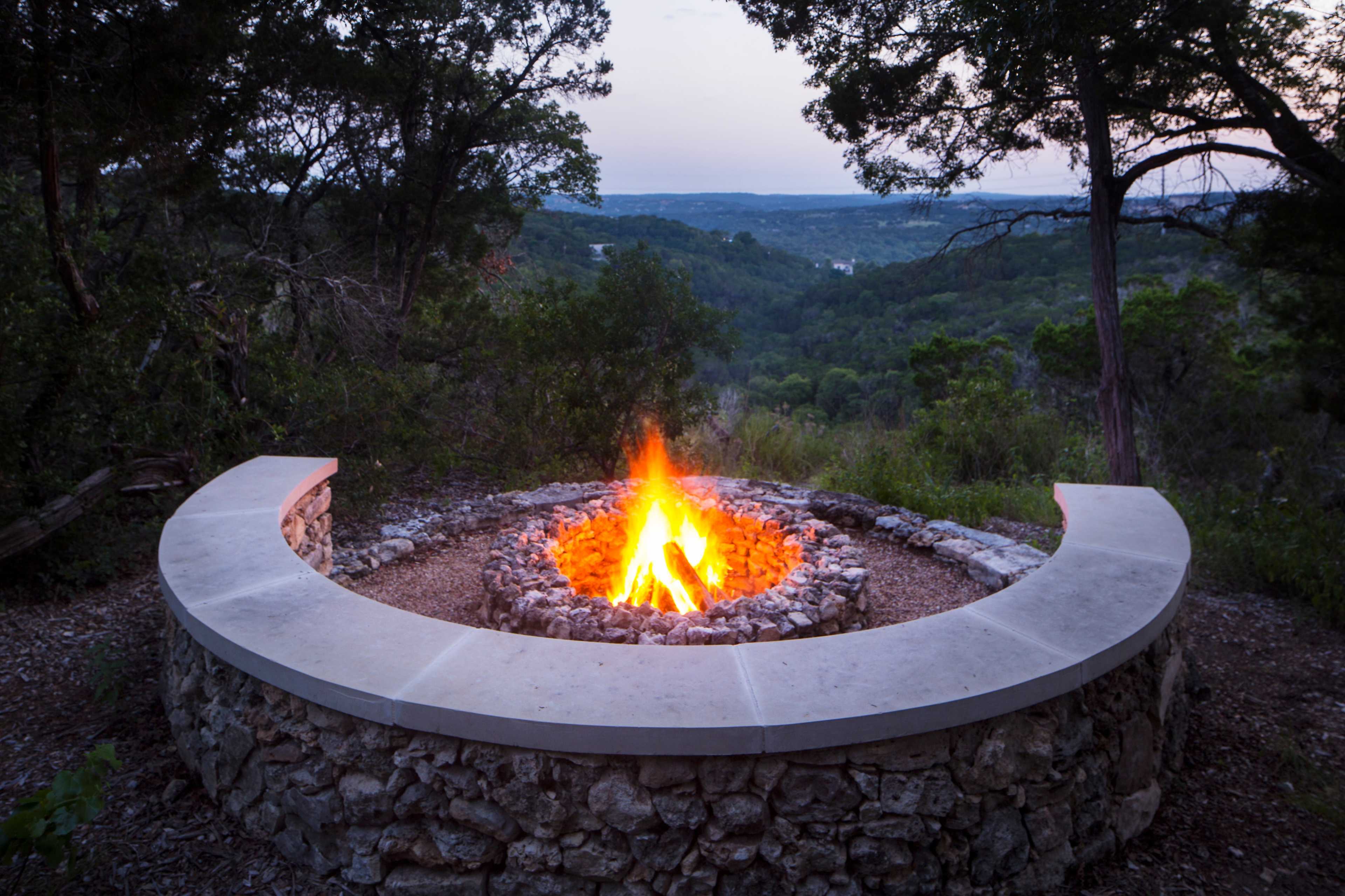 A circular stone fire pit with a vibrant flame is set against a backdrop of a lush, green hillside and a twilight sky.