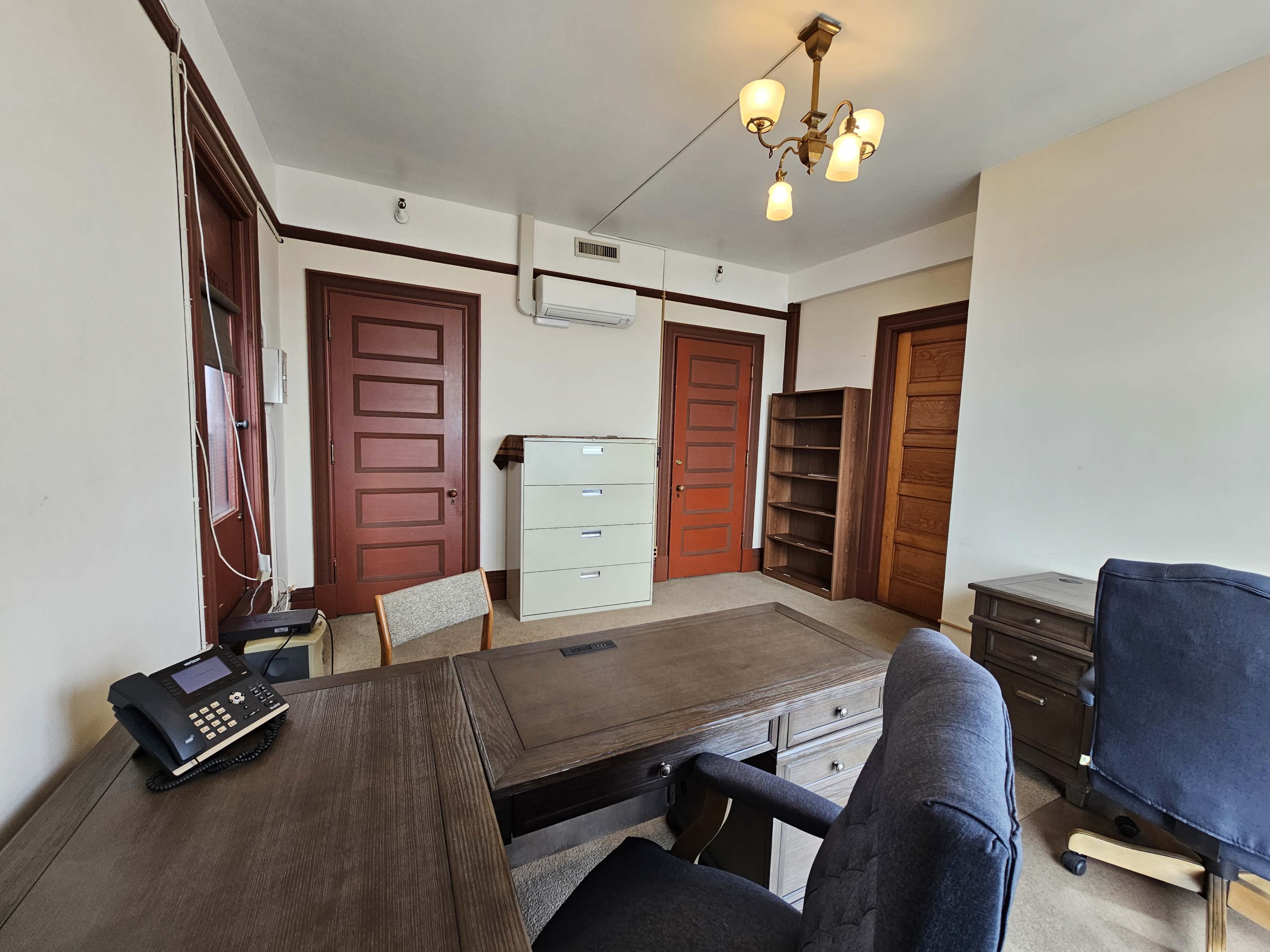 An office interior with a wooden desk, a chair, a filing cabinet, a bookcase, and several closed doors.