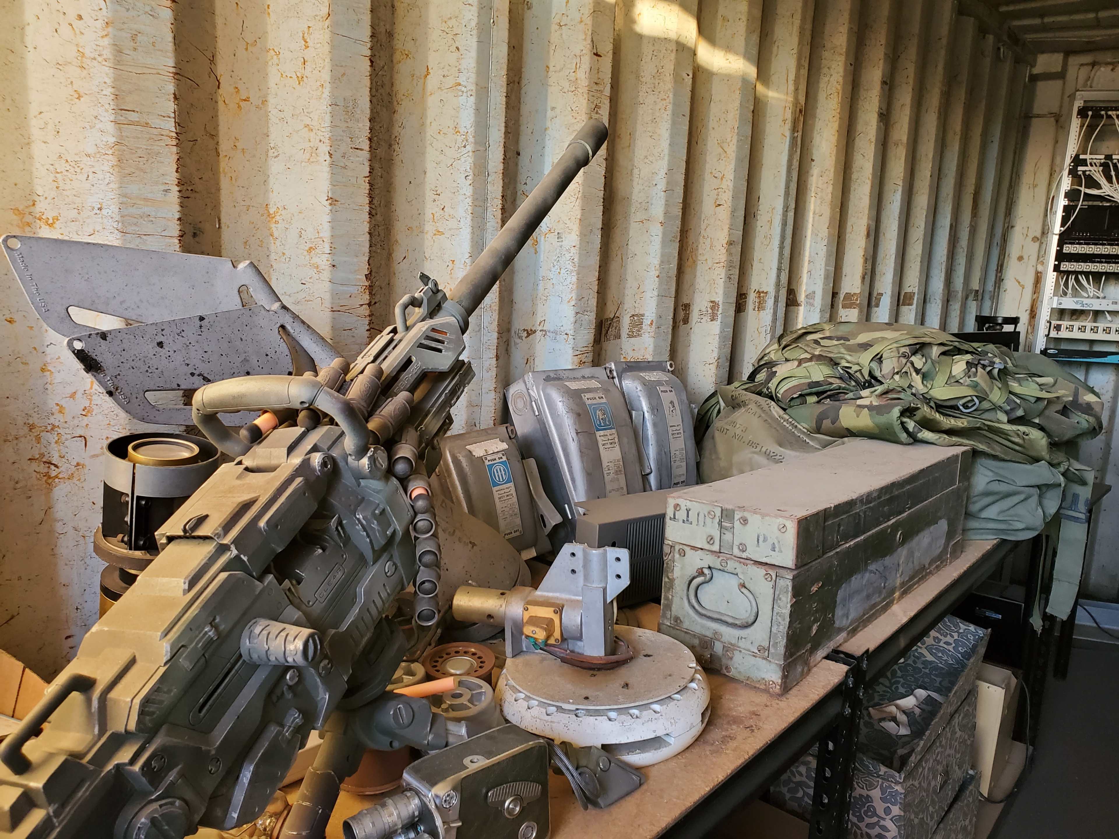 The image shows a collection of military equipment and gear, including a mounted weapon and various storage cases, arranged on a table inside a container.