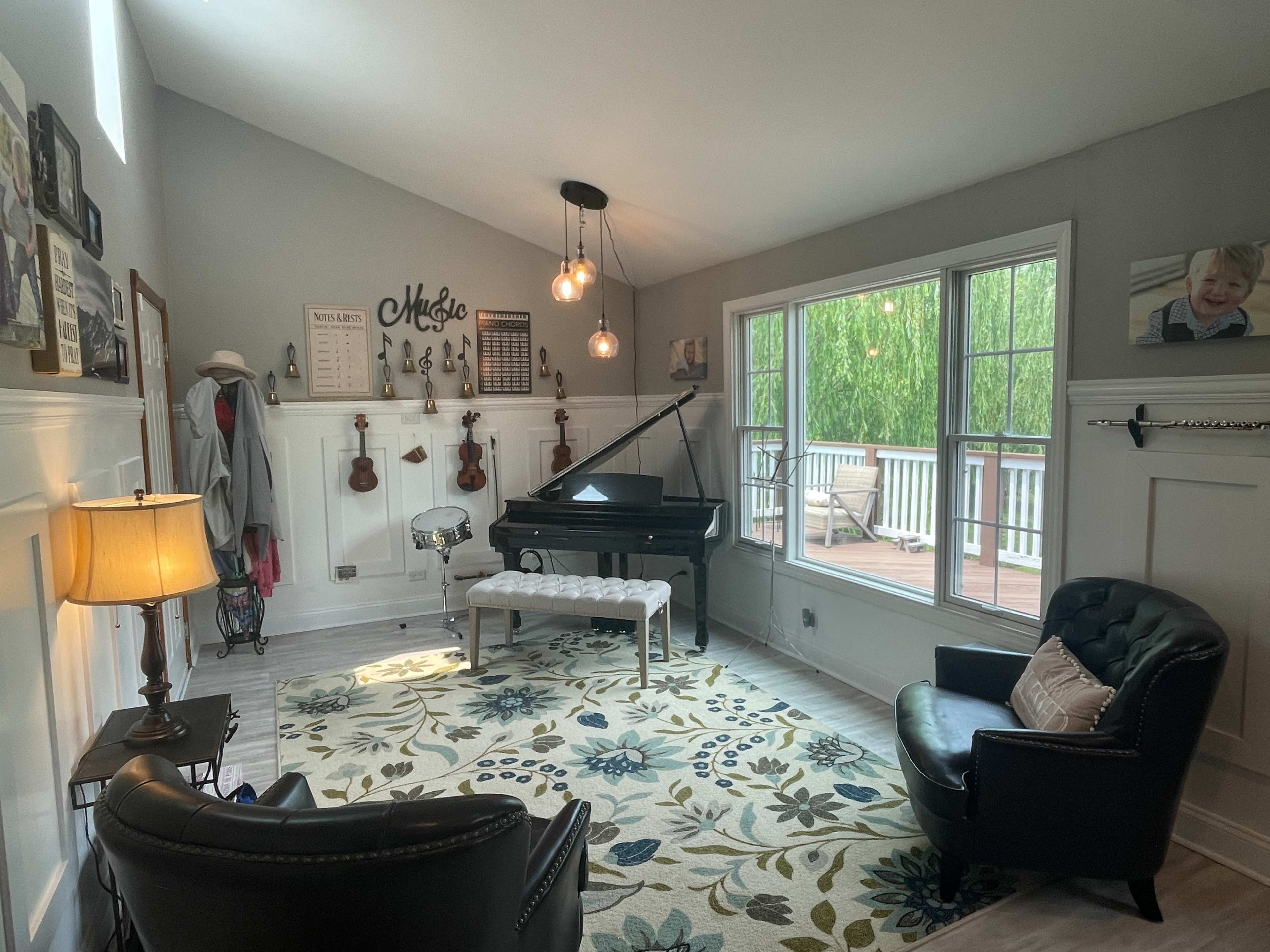 The room features a grand piano, two chairs, a decorative area rug, and large windows overlooking a wooden deck surrounded by greenery.
