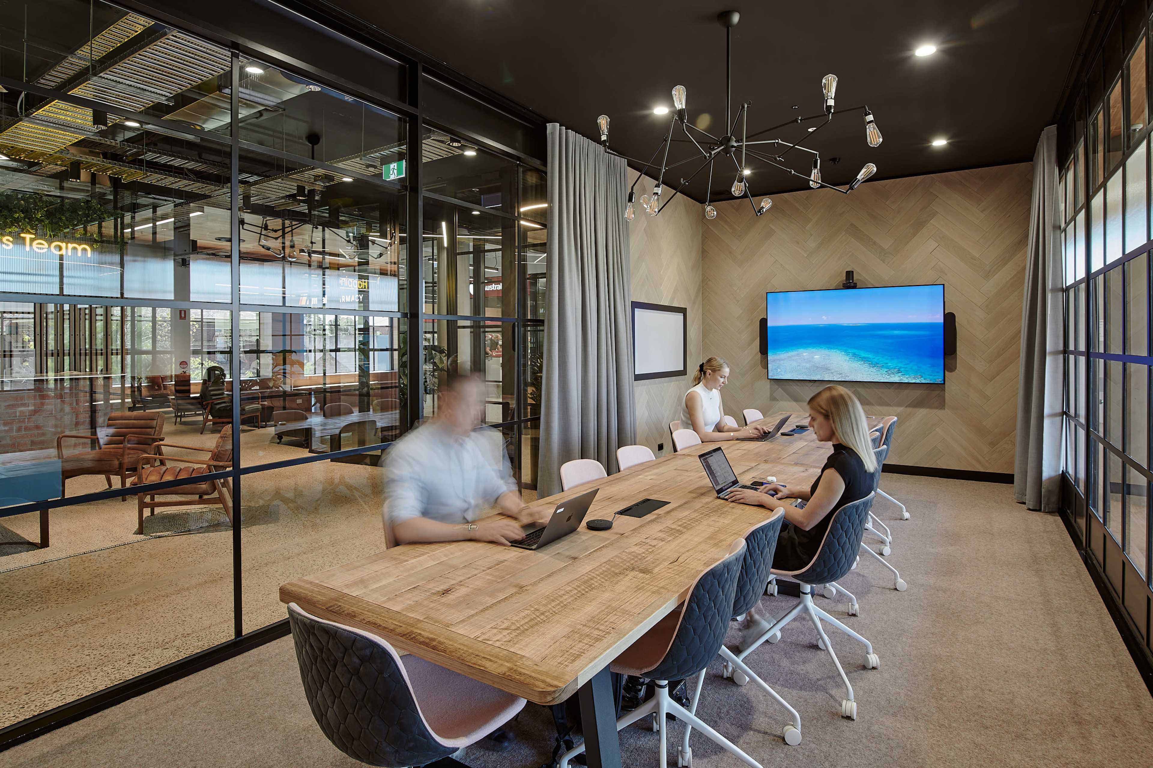 The image shows a modern office meeting room with a wooden conference table, three people working on laptops, and a large screen displaying a beach scene.