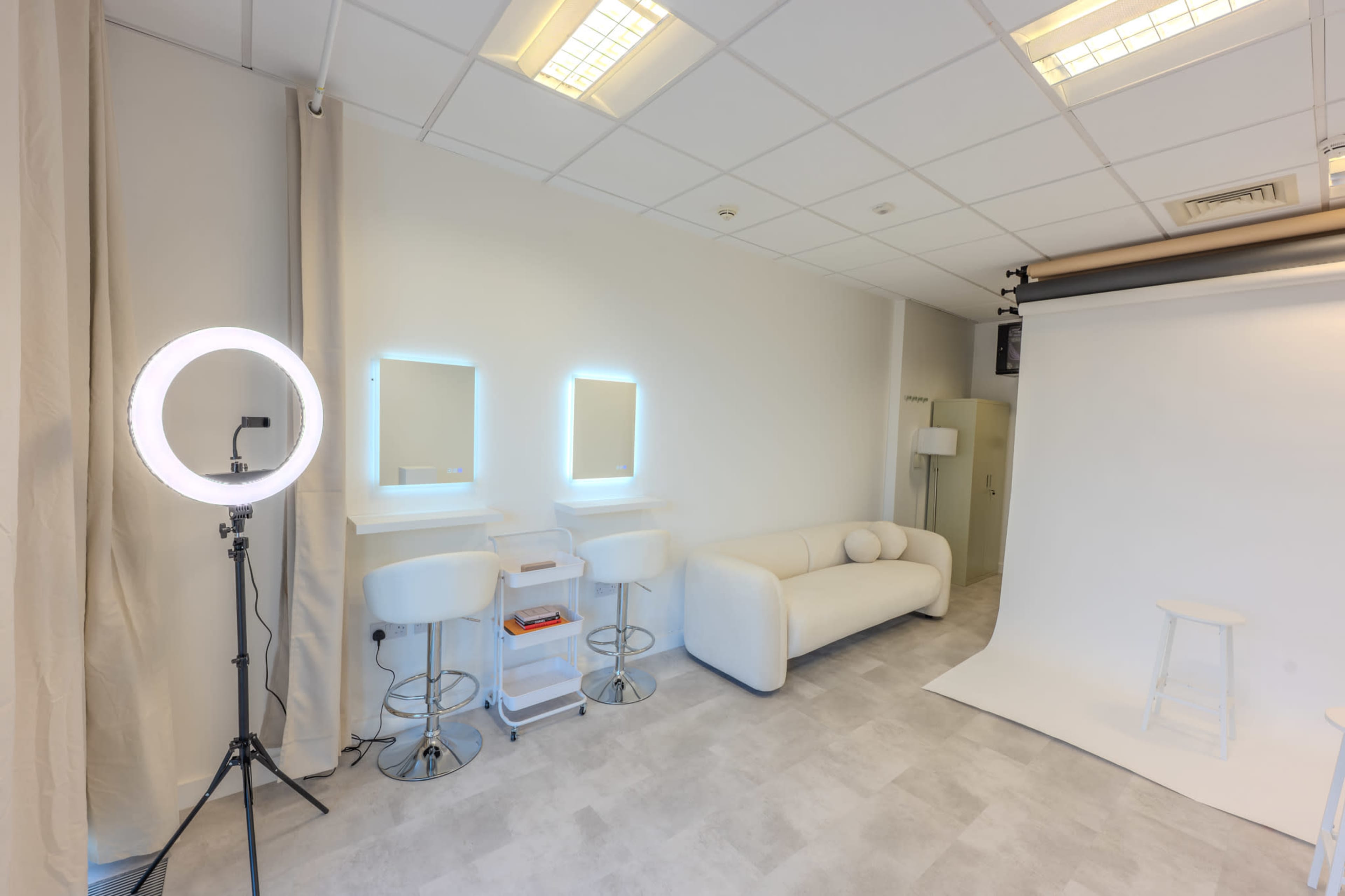 The image depicts a bright, minimalist photography studio featuring a white couch, tall bar stools, round mirrors, and a backdrop setup.