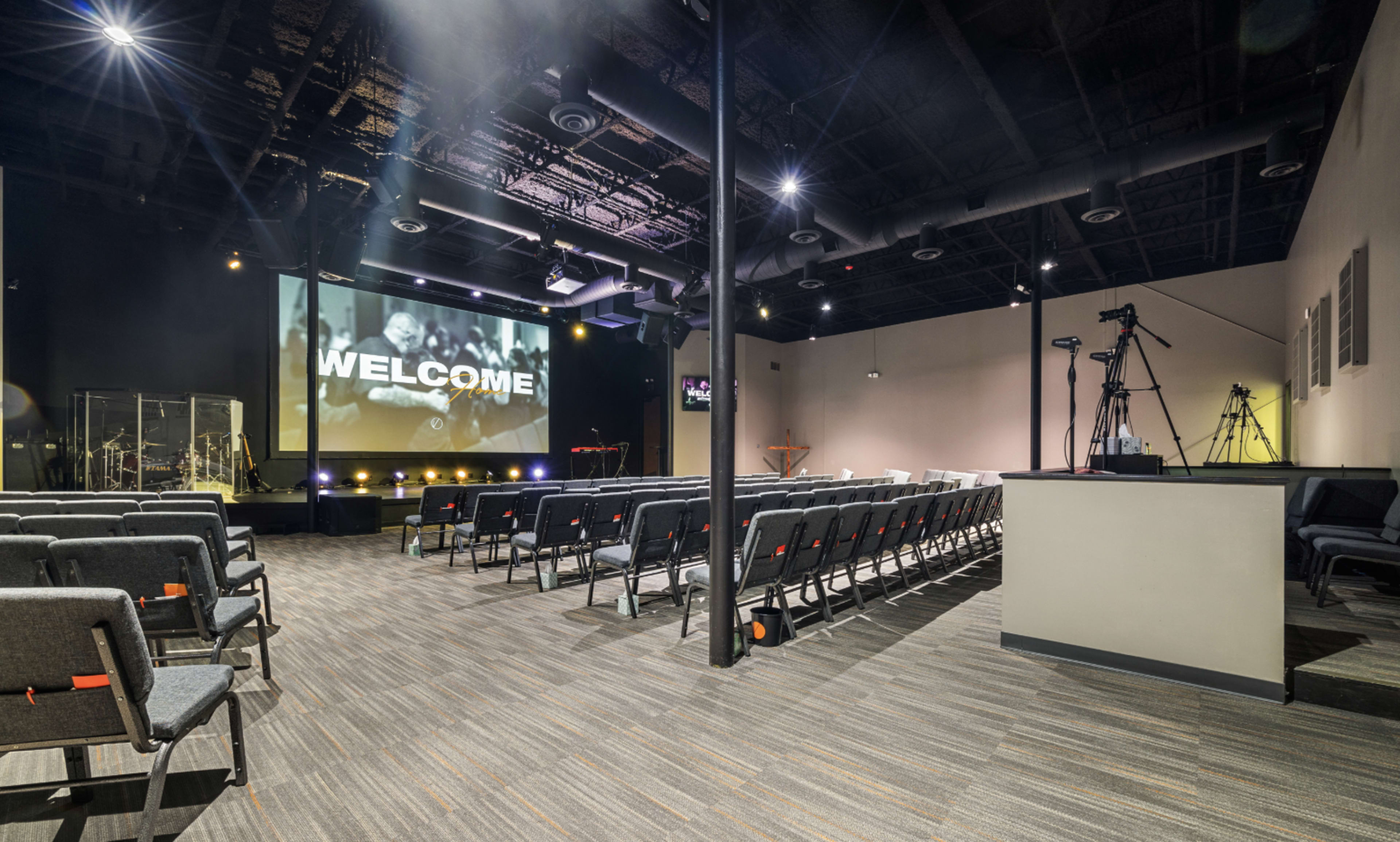 The image shows an interior view of a modern auditorium with rows of seats facing a large screen displaying the word "WELCOME," with stage lighting and equipment visible in the background.