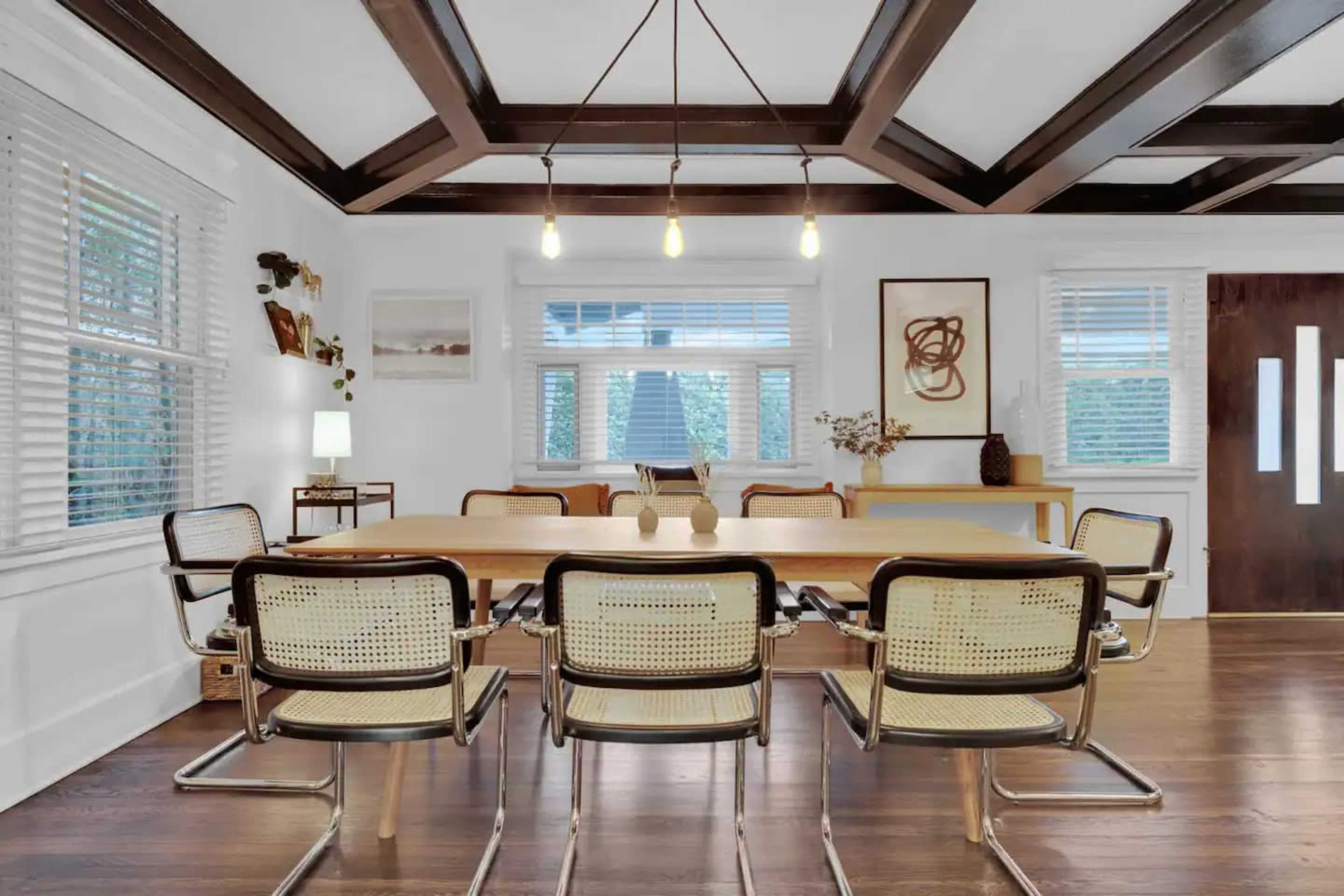 A dining room features a large wooden table surrounded by six metal and cane chairs, with a coffered ceiling and large windows allowing natural light to enter.