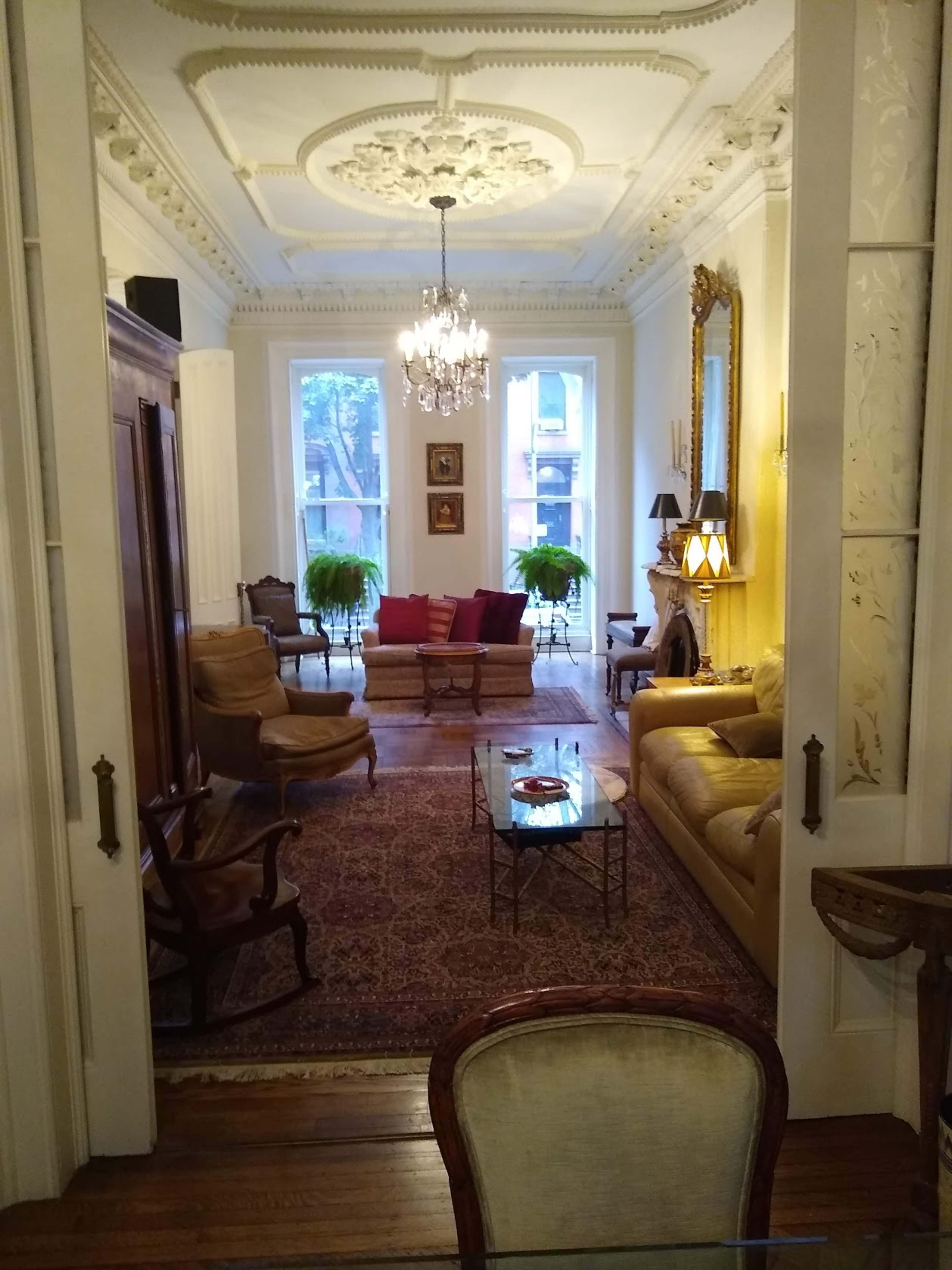 A stylish living room features a chandelier, ornate ceiling, and arranged seating around a coffee table, with large windows allowing natural light.