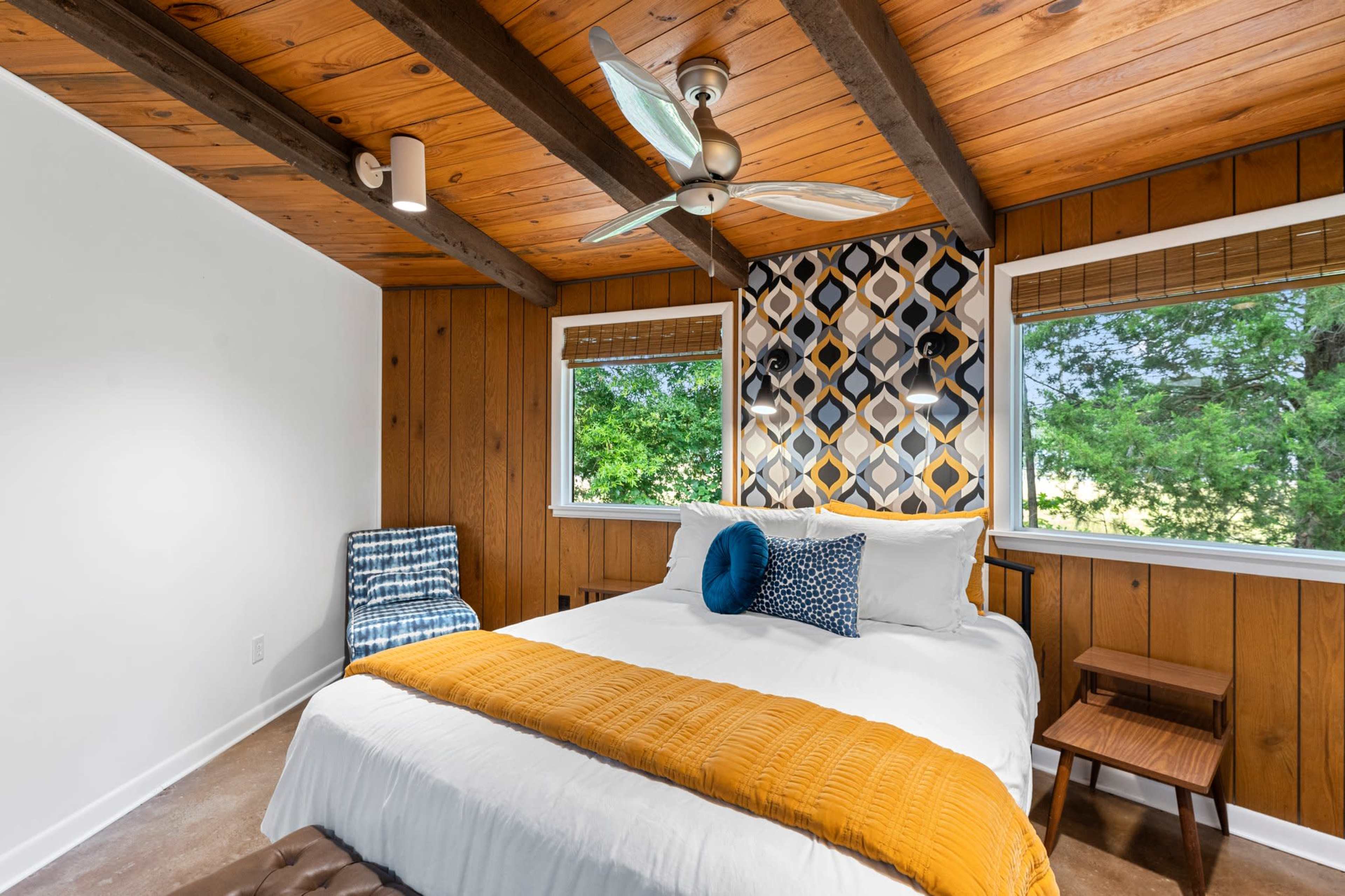 The image shows a bedroom with a bed adorned in white bedding and a mustard yellow throw, featuring wooden walls, a patterned accent wall, and large windows providing natural light.