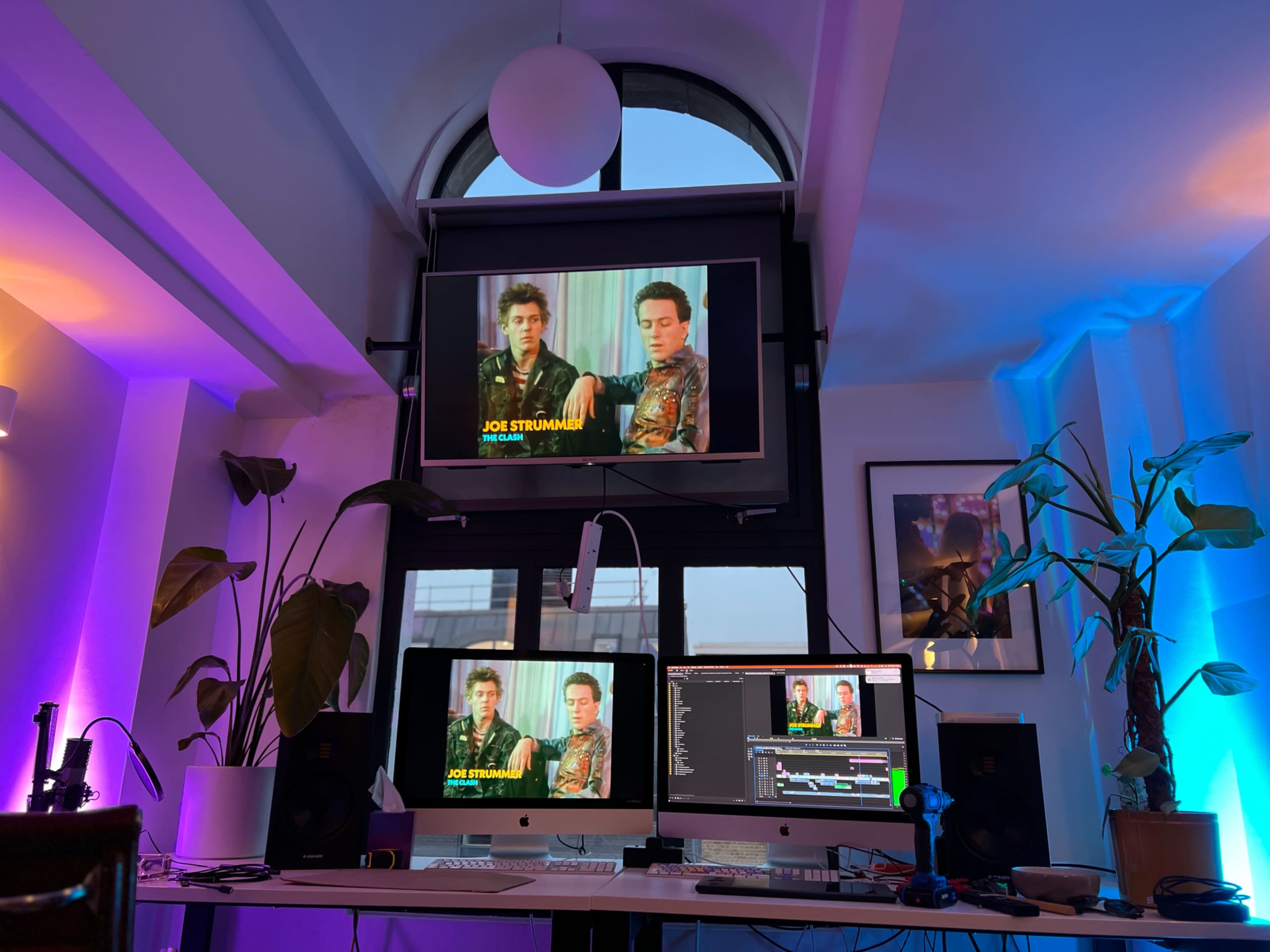 The image shows a workspace with three monitors displaying videos, a large window in the background, and plants placed near the desk.