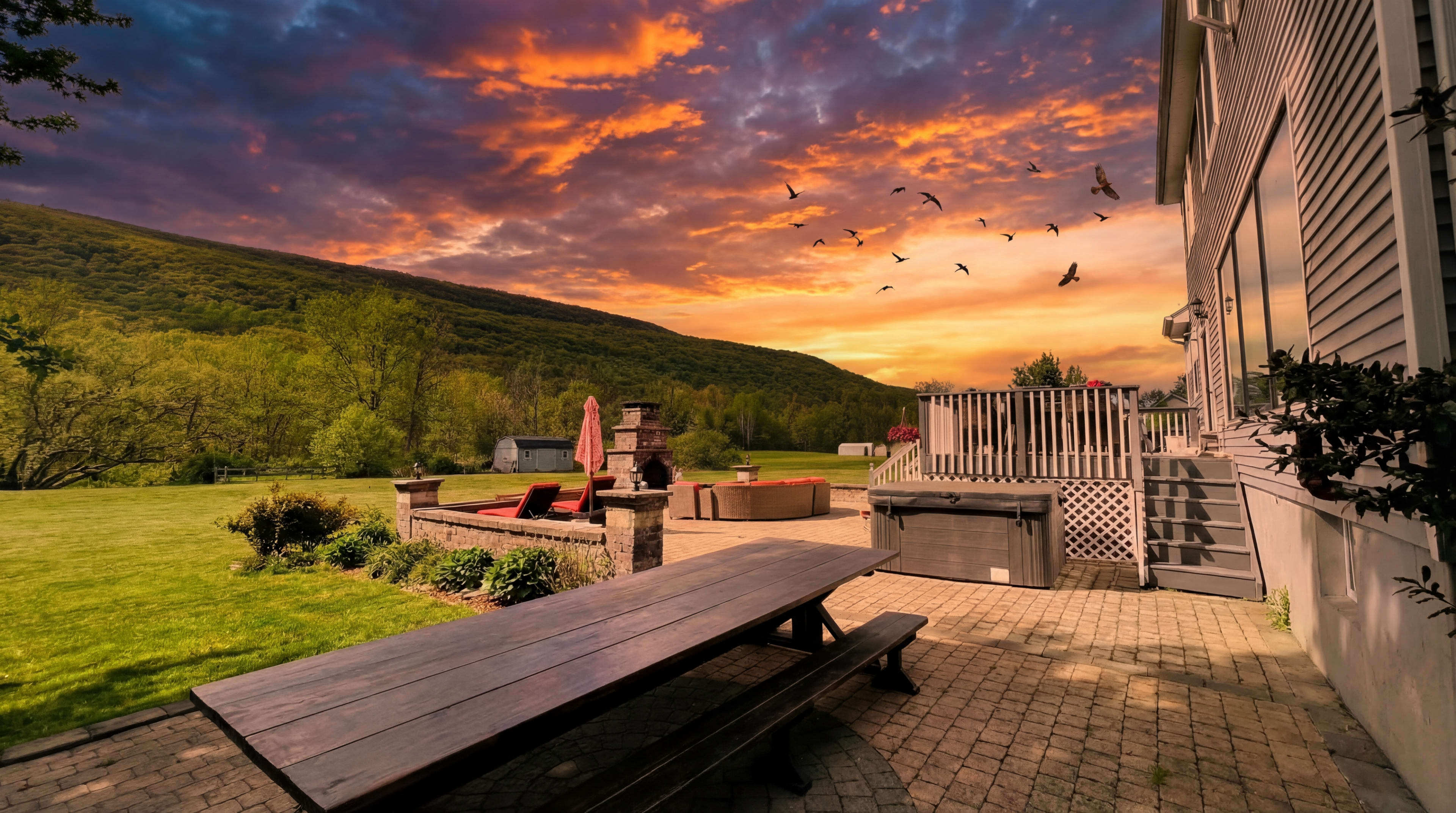 A long wooden table sits on a brick patio overlooking a grassy yard and a mountain range under a colorful sunset sky with birds flying overhead.