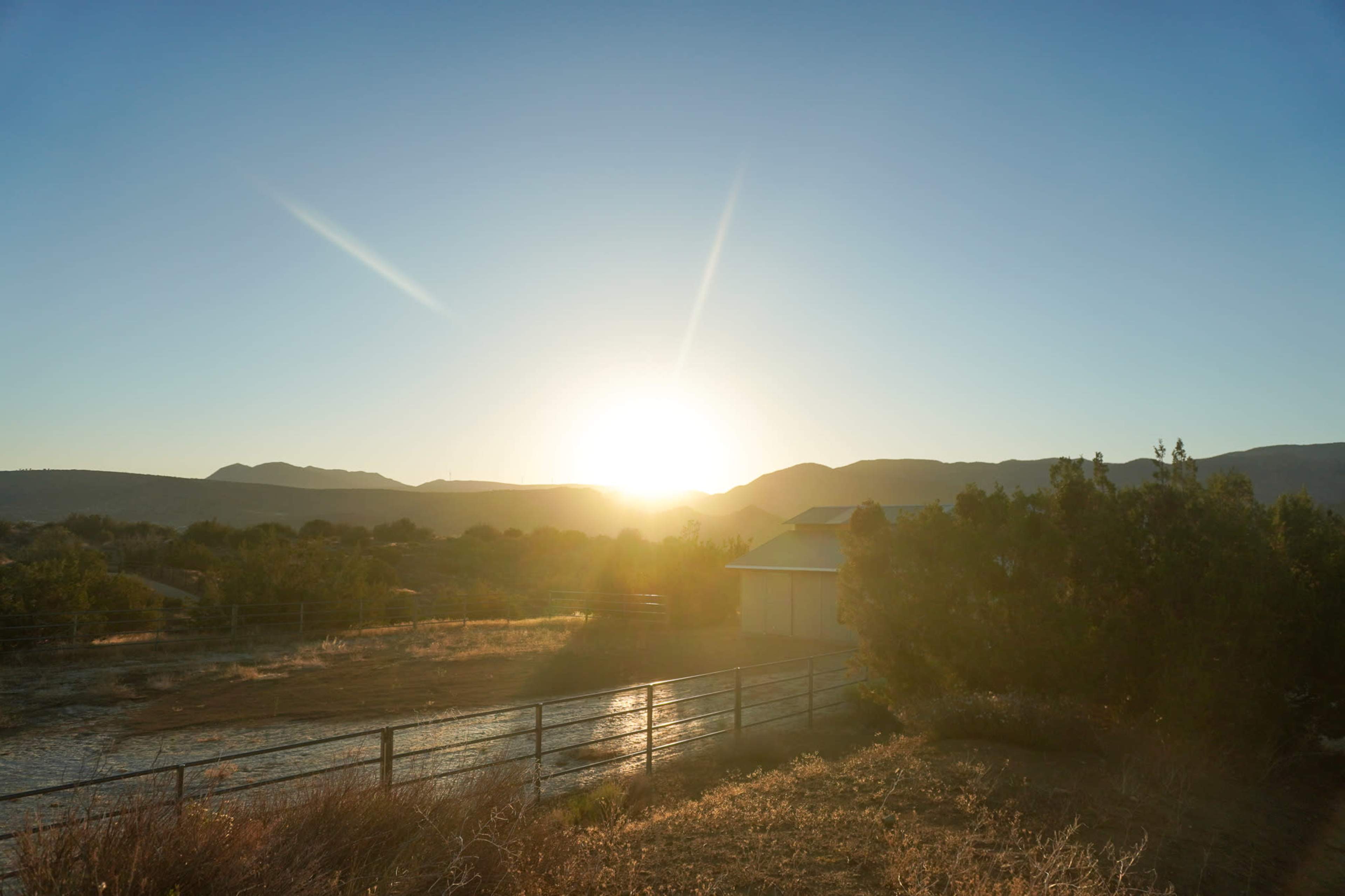 The sun rises over distant mountains, illuminating a quiet landscape with a fence and a building in the foreground.