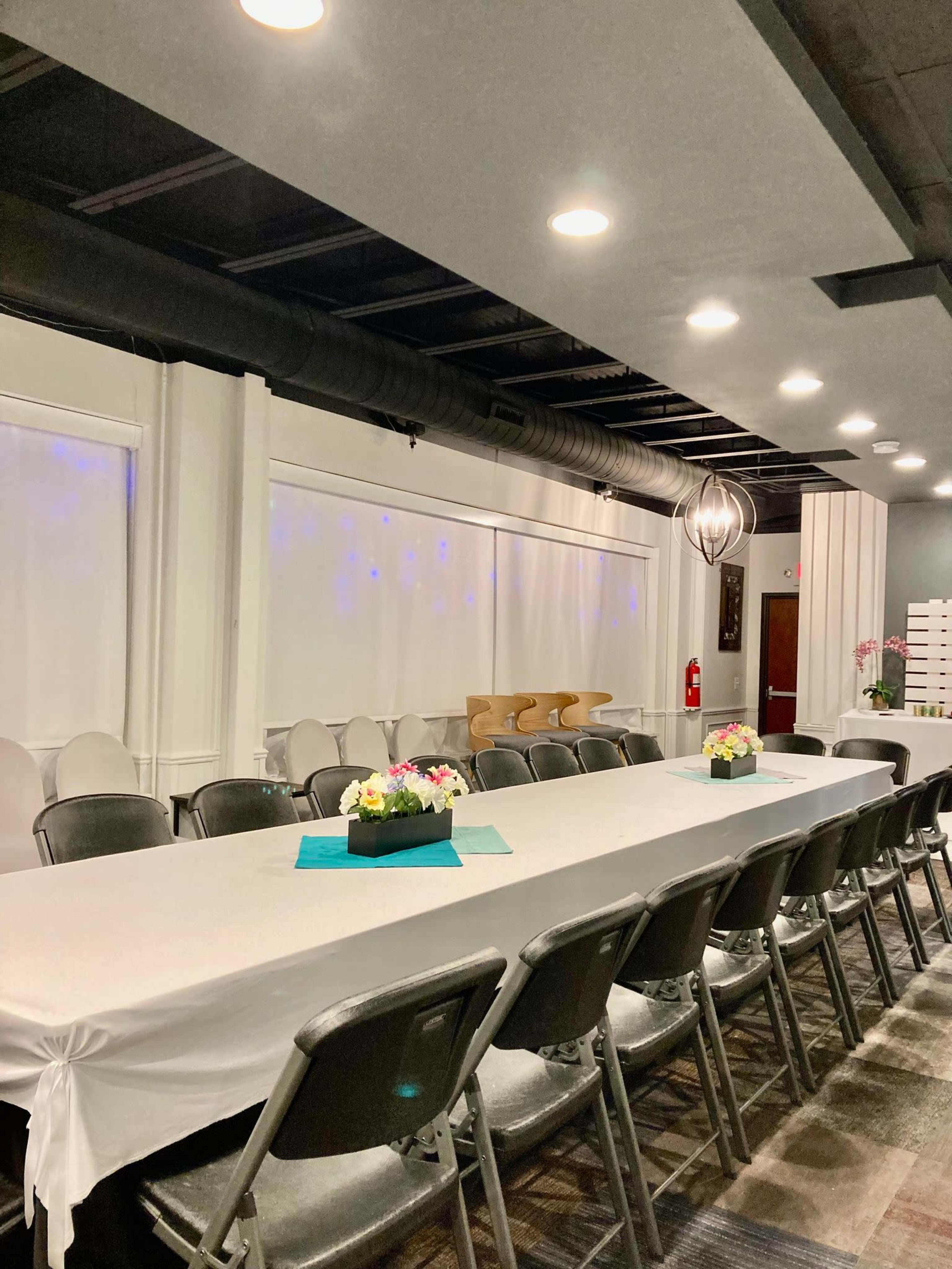 A spacious event room with a long table covered in a white tablecloth, surrounded by silver chairs, and adorned with flower arrangements in the center.