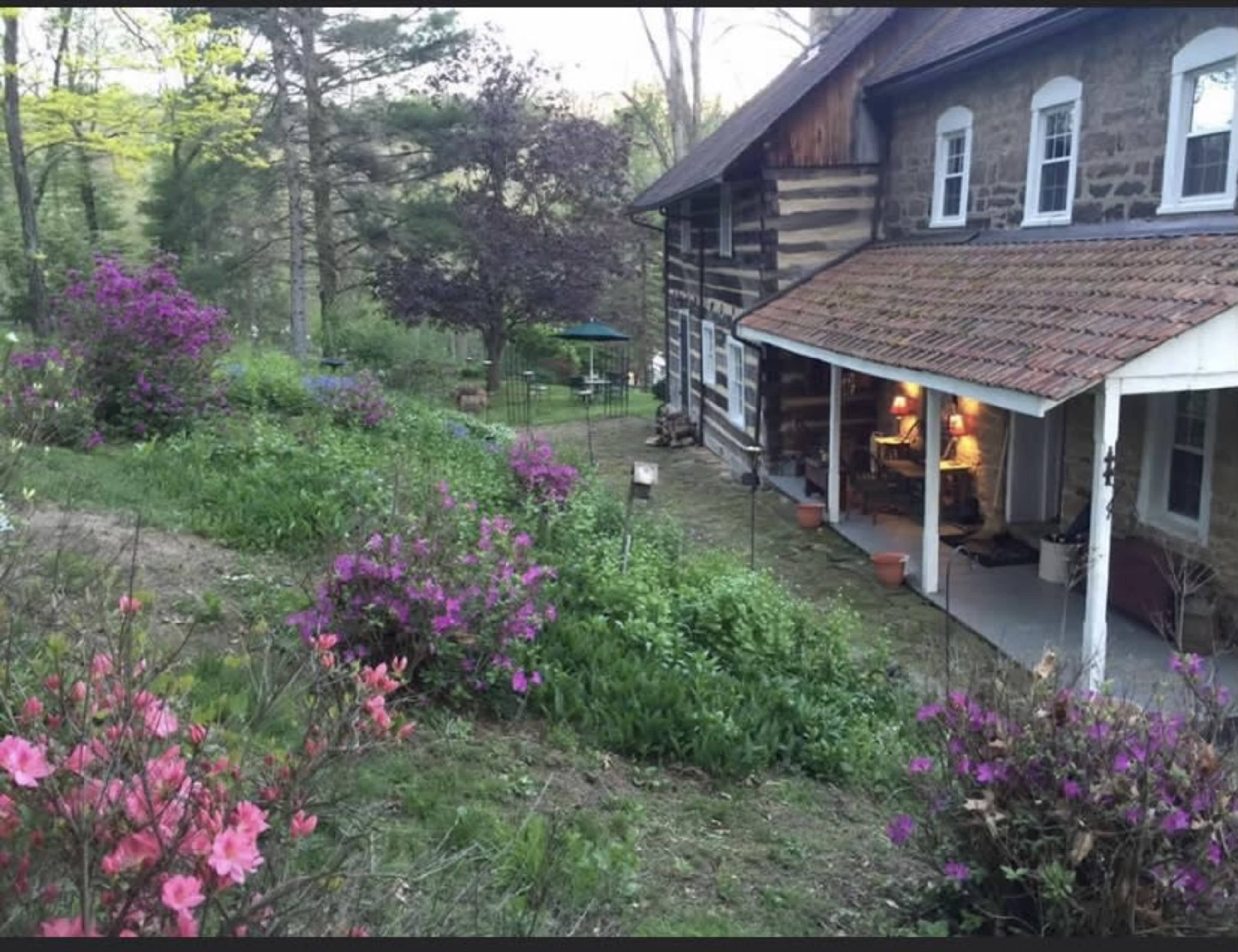 A stone house with a wooden porch is surrounded by blooming azaleas and a garden in a wooded area.