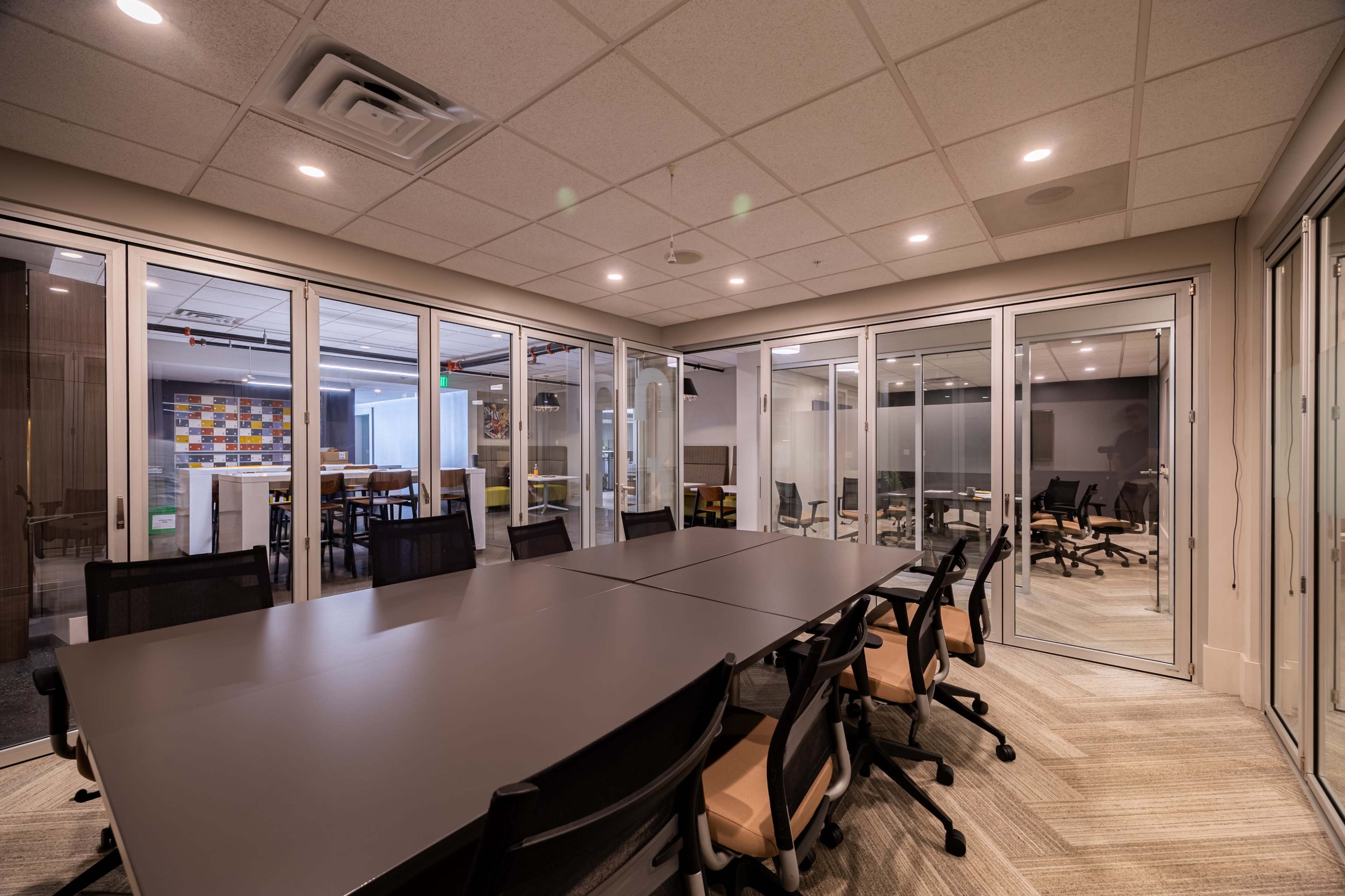The image shows a modern conference room with a large table surrounded by black and beige chairs, featuring glass walls and a view into an adjacent space.