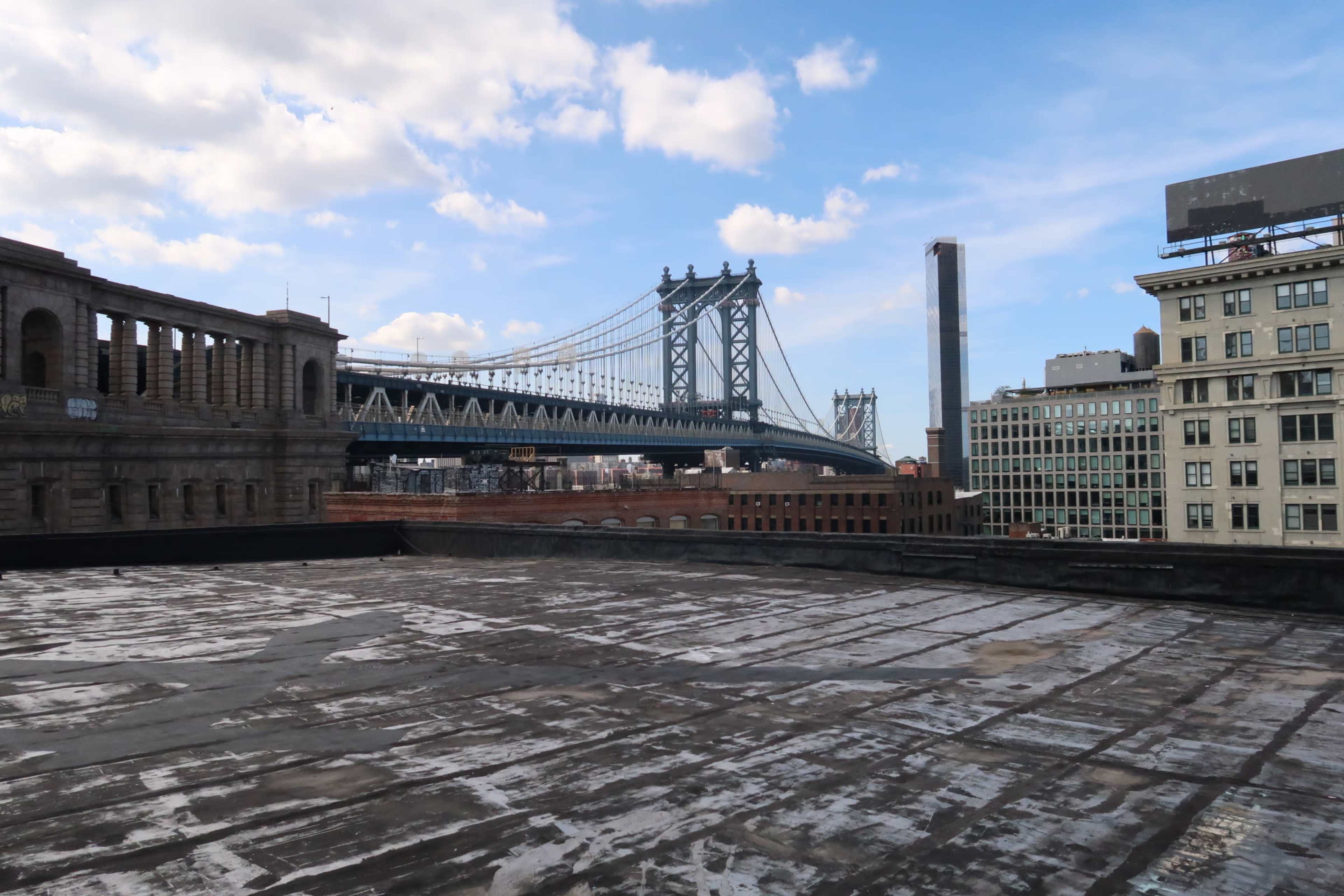 A rooftop view of the Manhattan Bridge with buildings in the background under a partly cloudy sky.