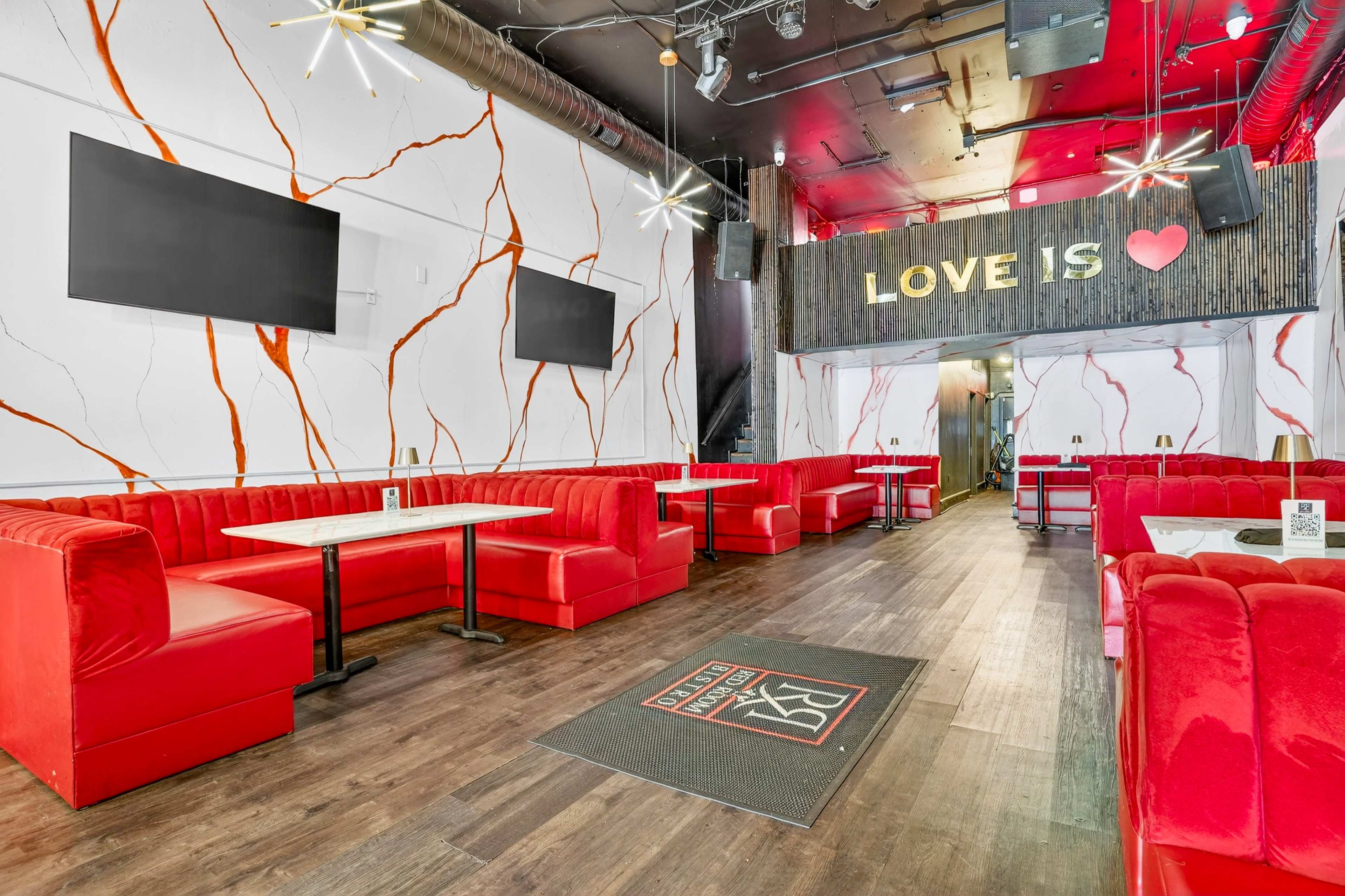The image shows a modern restaurant interior featuring red booth seating, a marbled wall design, and two large flat-screen televisions, with the words "LOVE IS" displayed above a stage area.