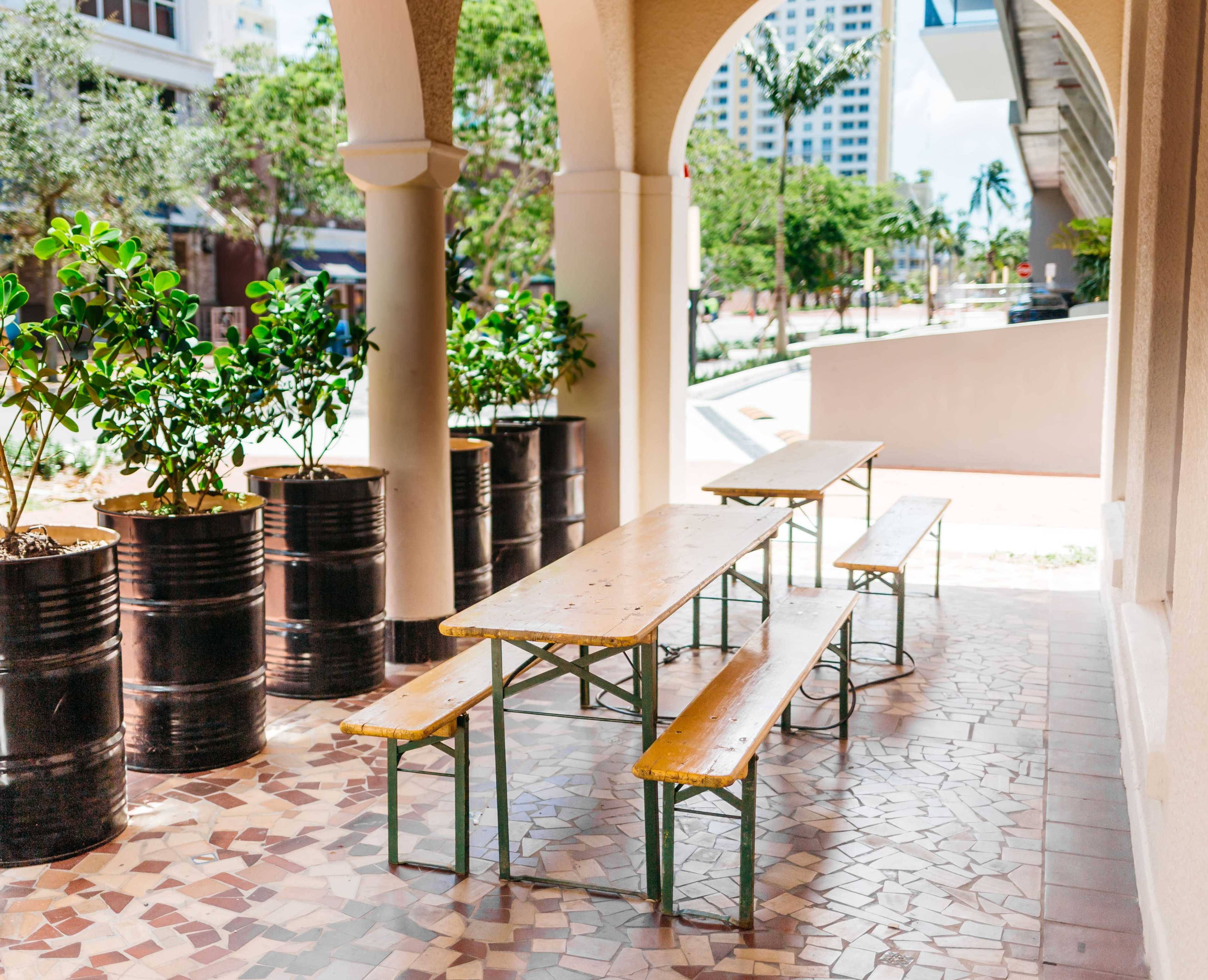 A courtyard area with wooden tables and benches, surrounded by potted plants and flanked by arched columns.