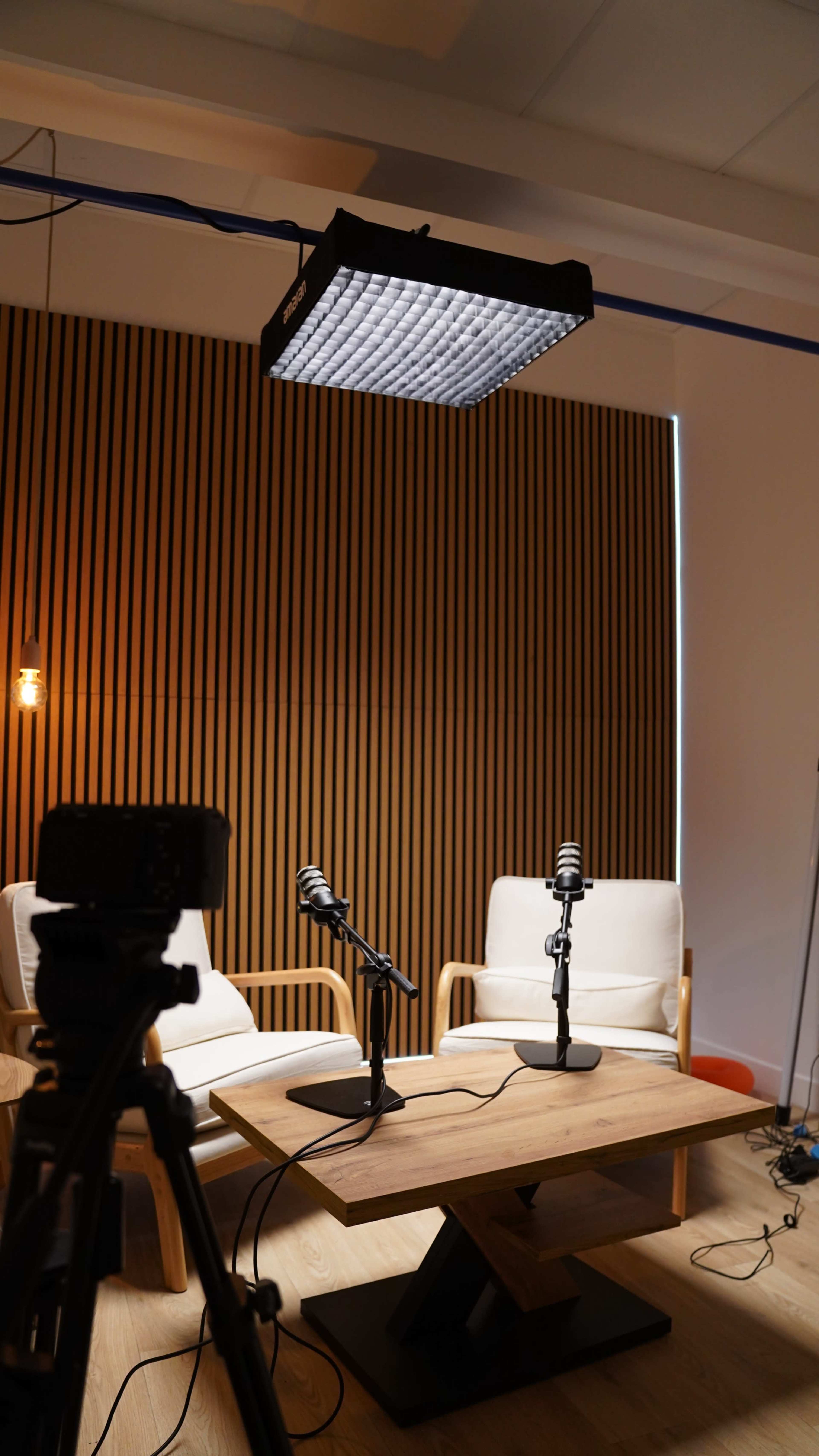 The image shows a modern podcast recording studio with two white chairs, two microphones on stands, and a wooden table, set against a backdrop of vertical wooden paneling and soft lighting.