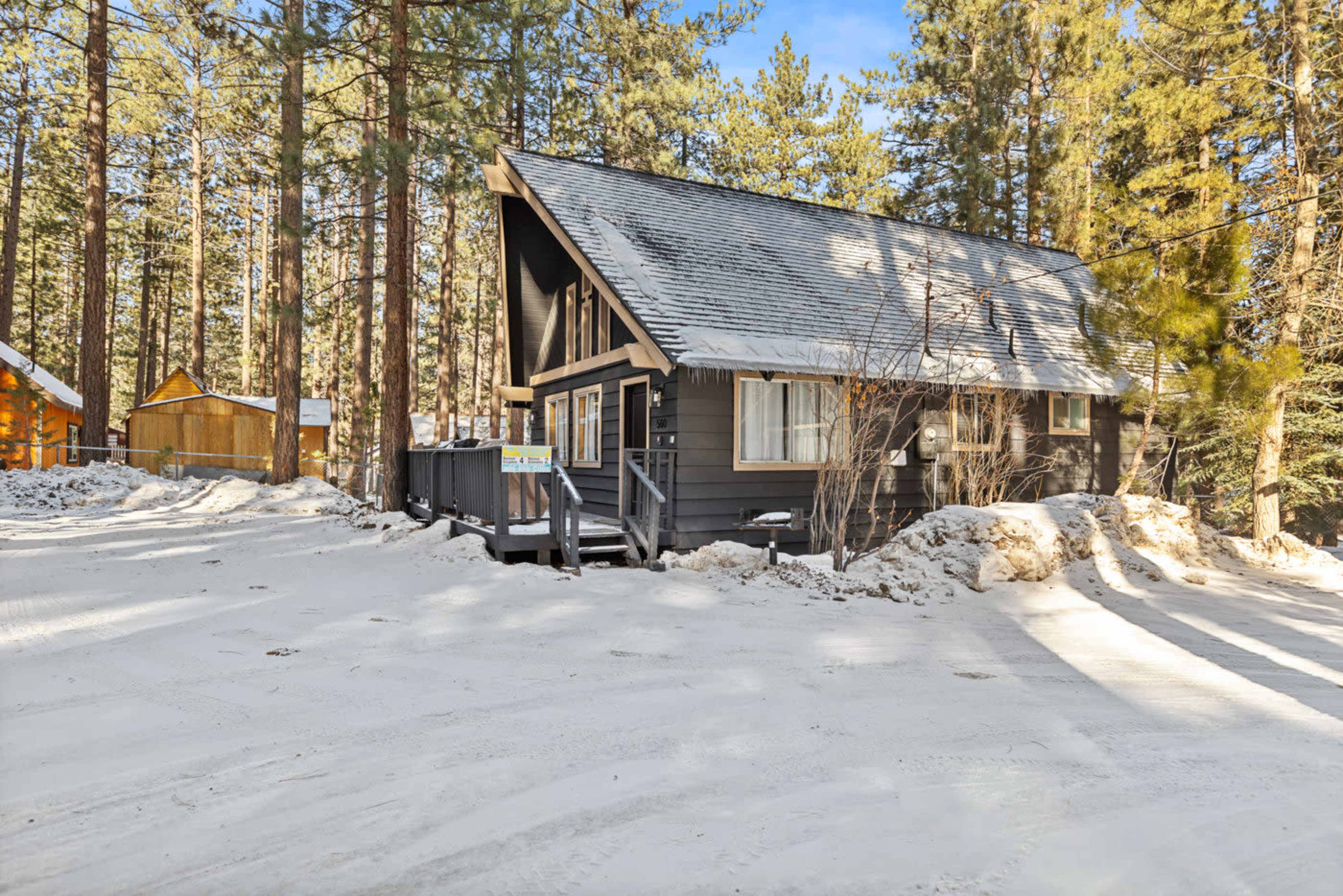 A chalet-style house with a sloped roof is set among tall pine trees and surrounded by patches of snow.