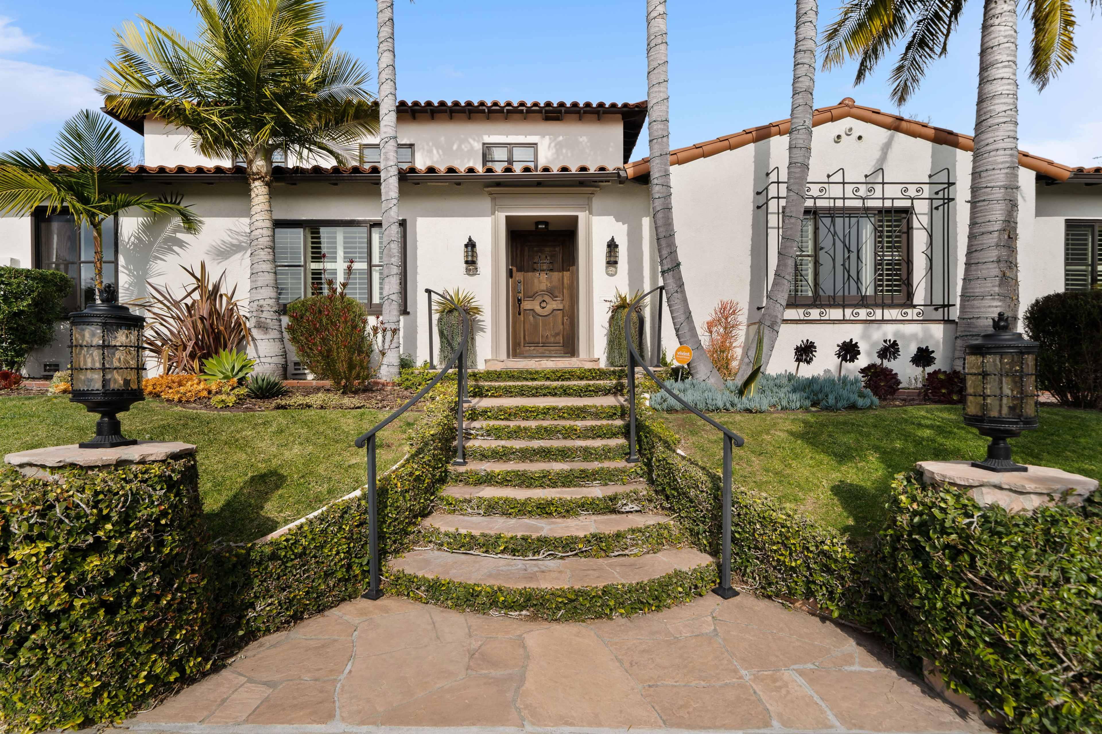 The image shows a two-story, Mediterranean-style house with a stone pathway leading to the front door, surrounded by well-manicured lawns and palm trees.