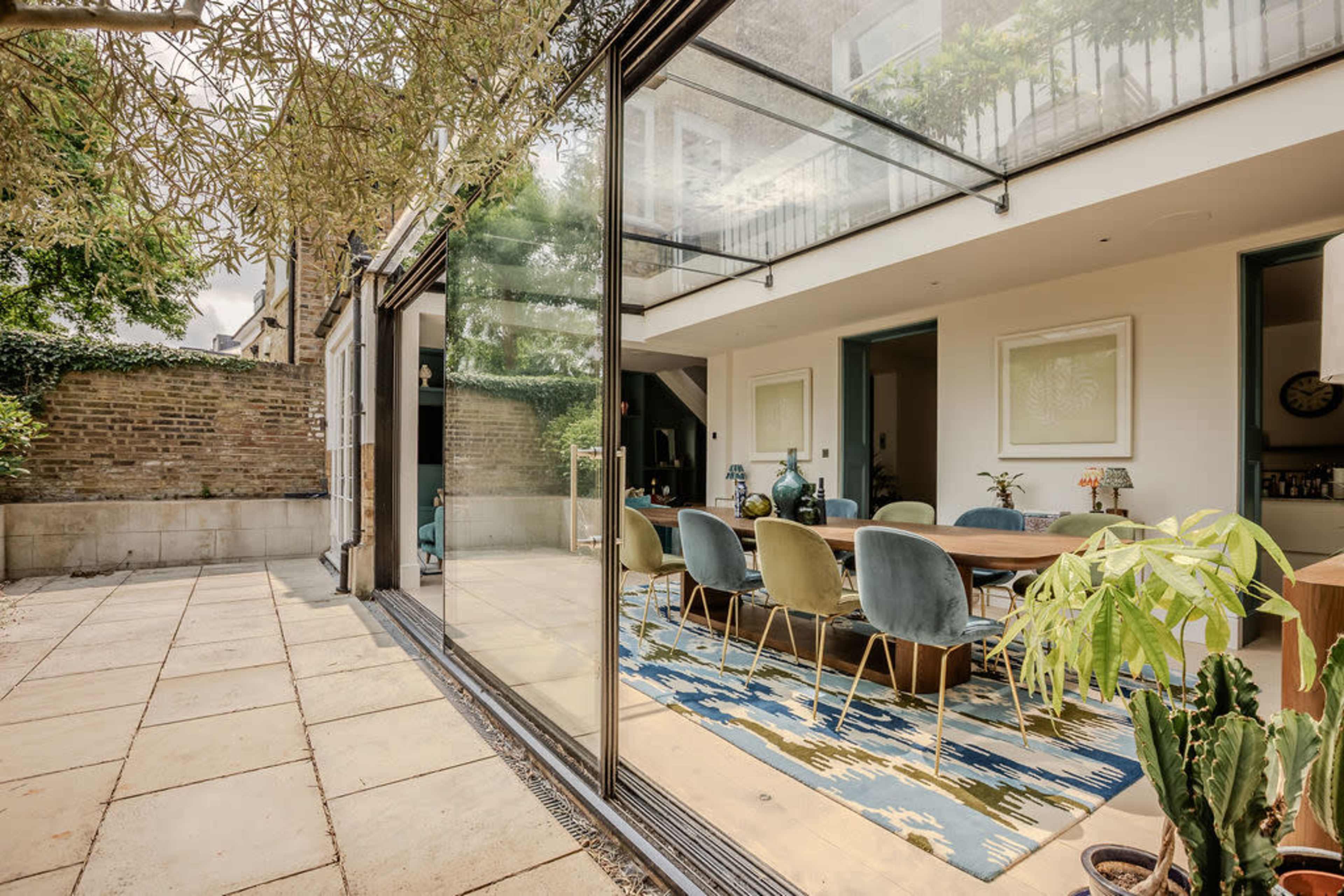 The image shows a modern outdoor patio with a sliding glass door leading to an interior dining area featuring a long table and colorful chairs.