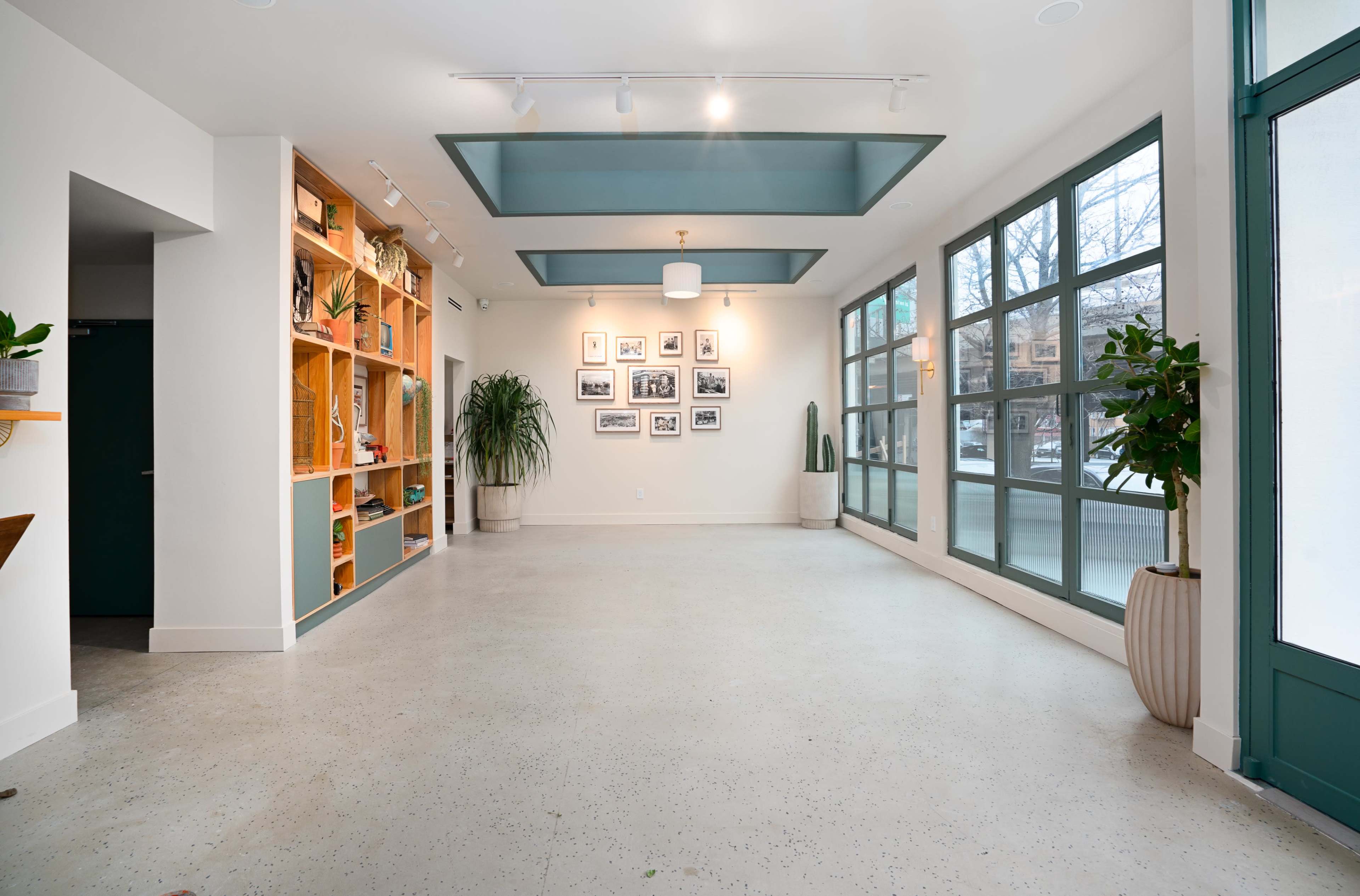 A spacious, well-lit room features large windows, a concrete floor, and wooden shelves filled with plants and decorative items.