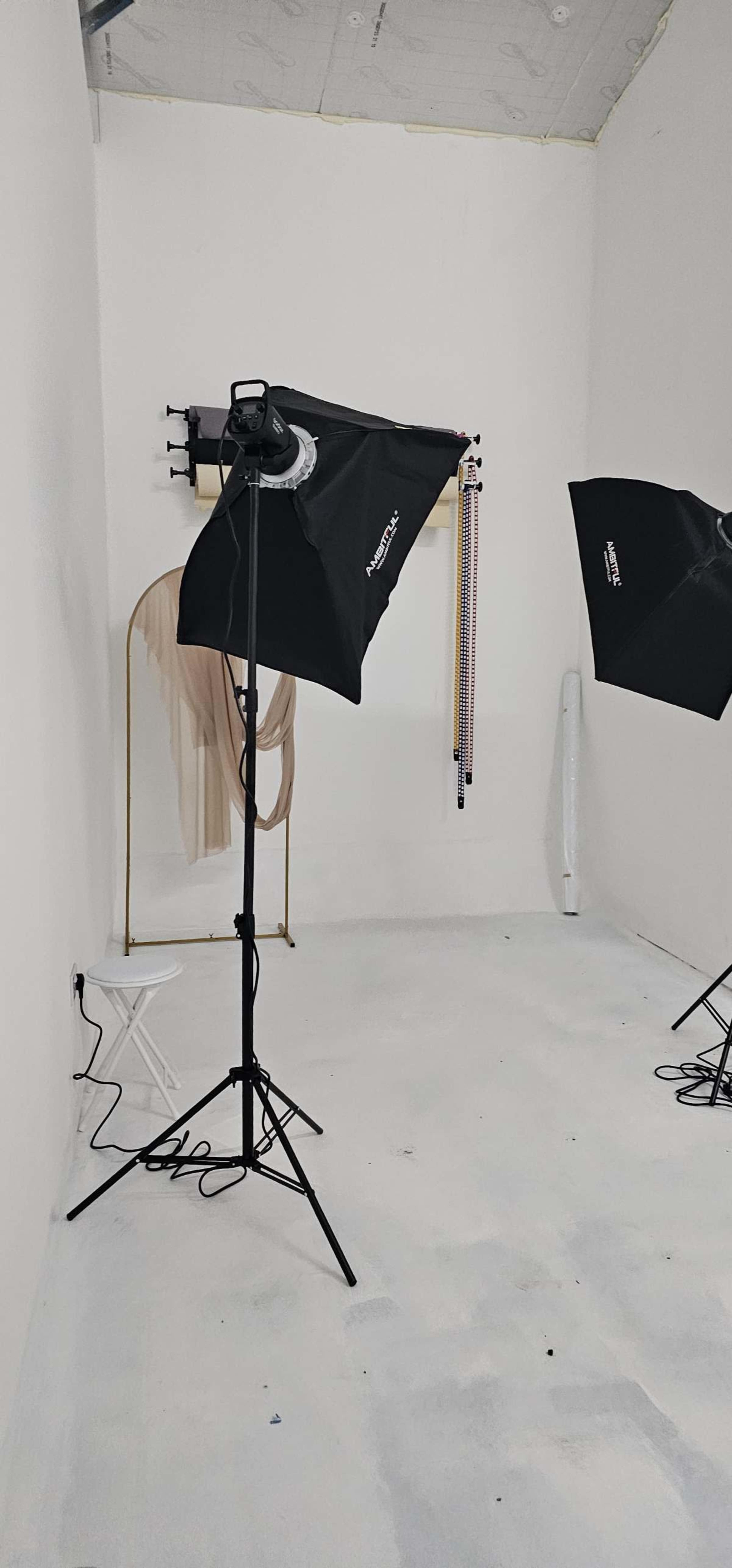 The image shows a photography studio with two softbox lights, a clothing rack with hanging garments, and a small folding chair against a white wall.