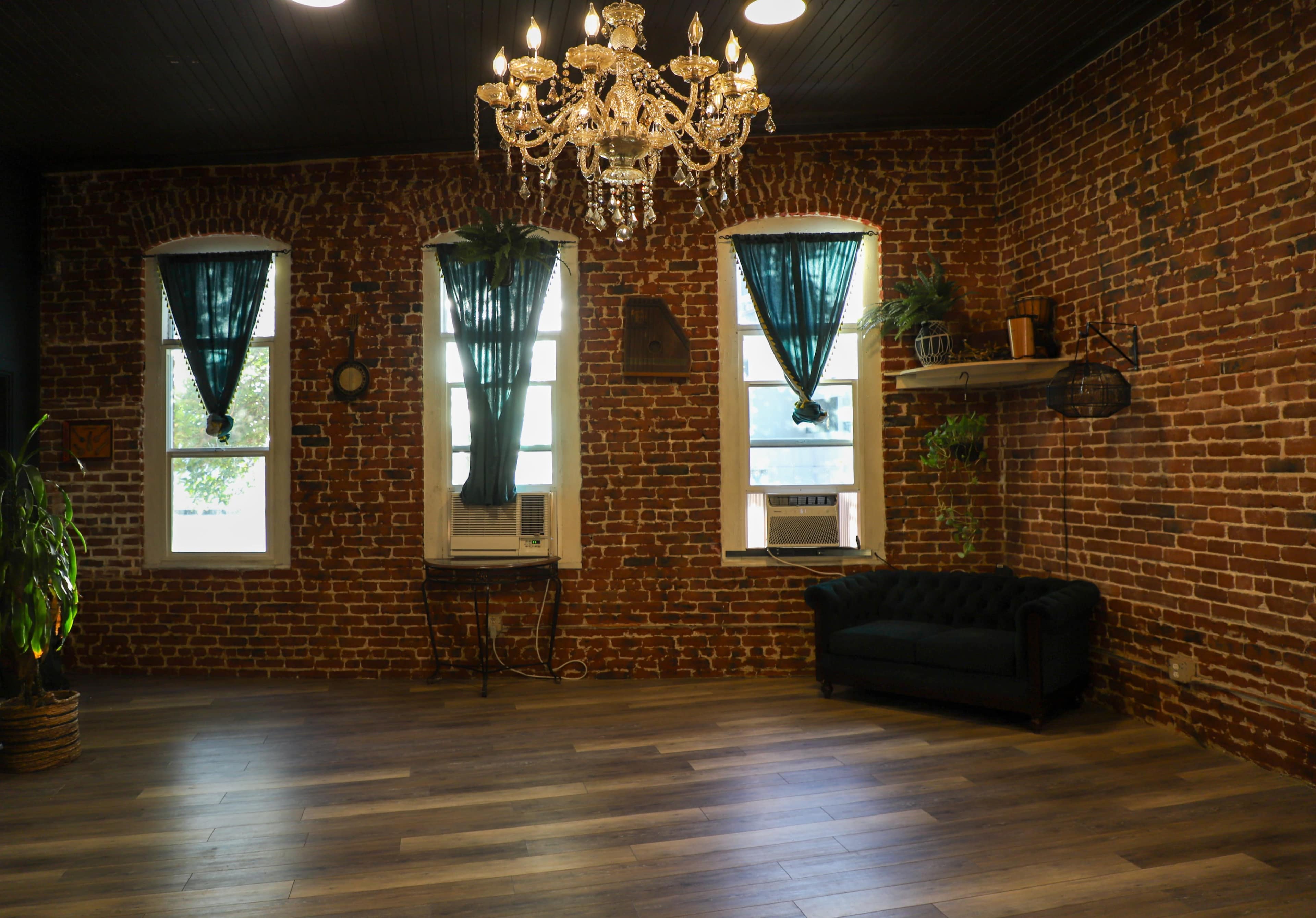 The scene features a room with exposed brick walls, large windows dressed with teal curtains, a chandelier overhead, and a dark couch positioned on hardwood flooring.