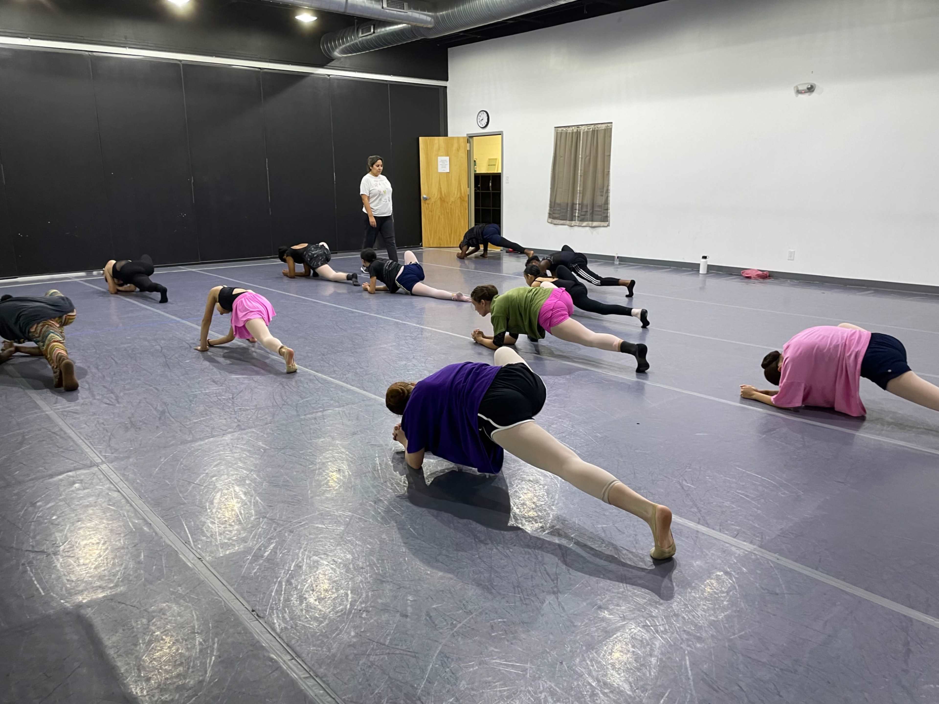 A group of dancers stretches on the floor of a rehearsal studio while an instructor observes in the background.