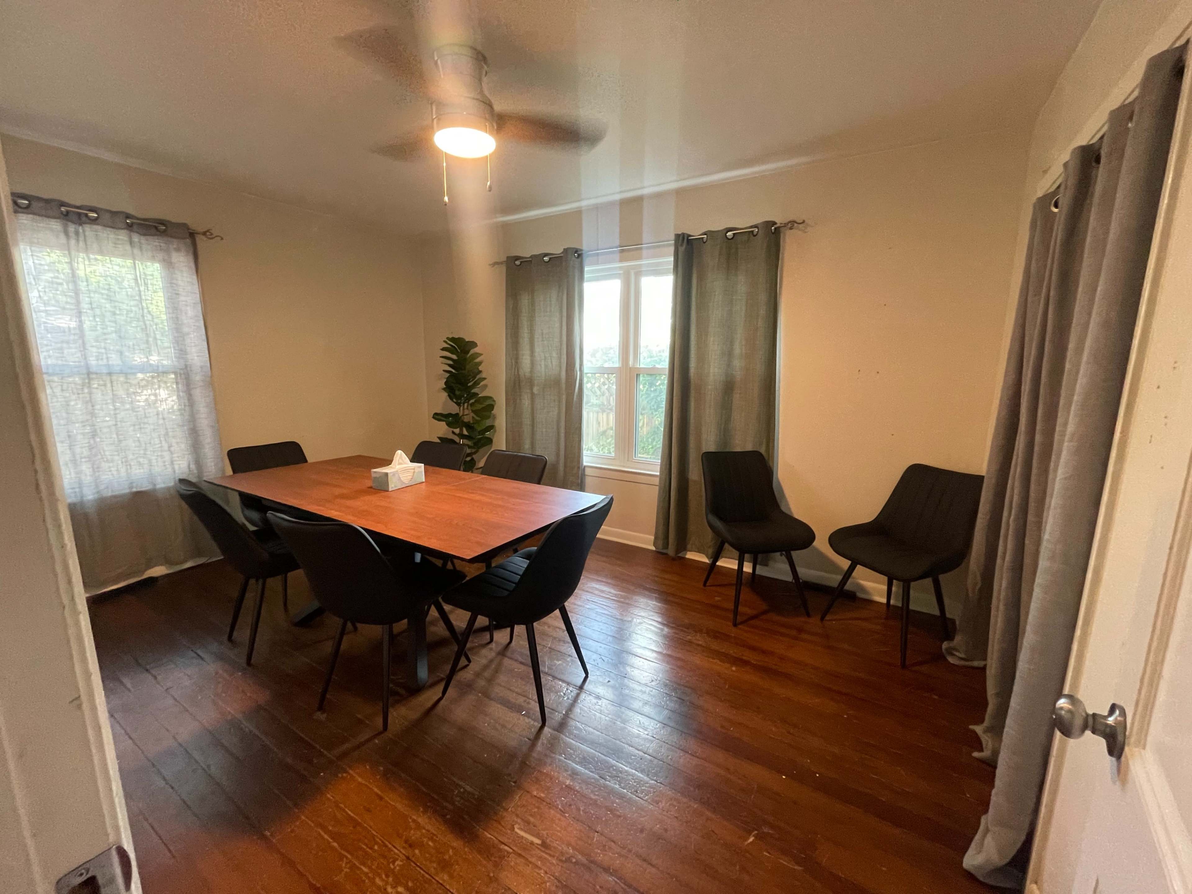 A dining room features a wooden table surrounded by black chairs, with natural light coming through a window covered by sheer curtains.