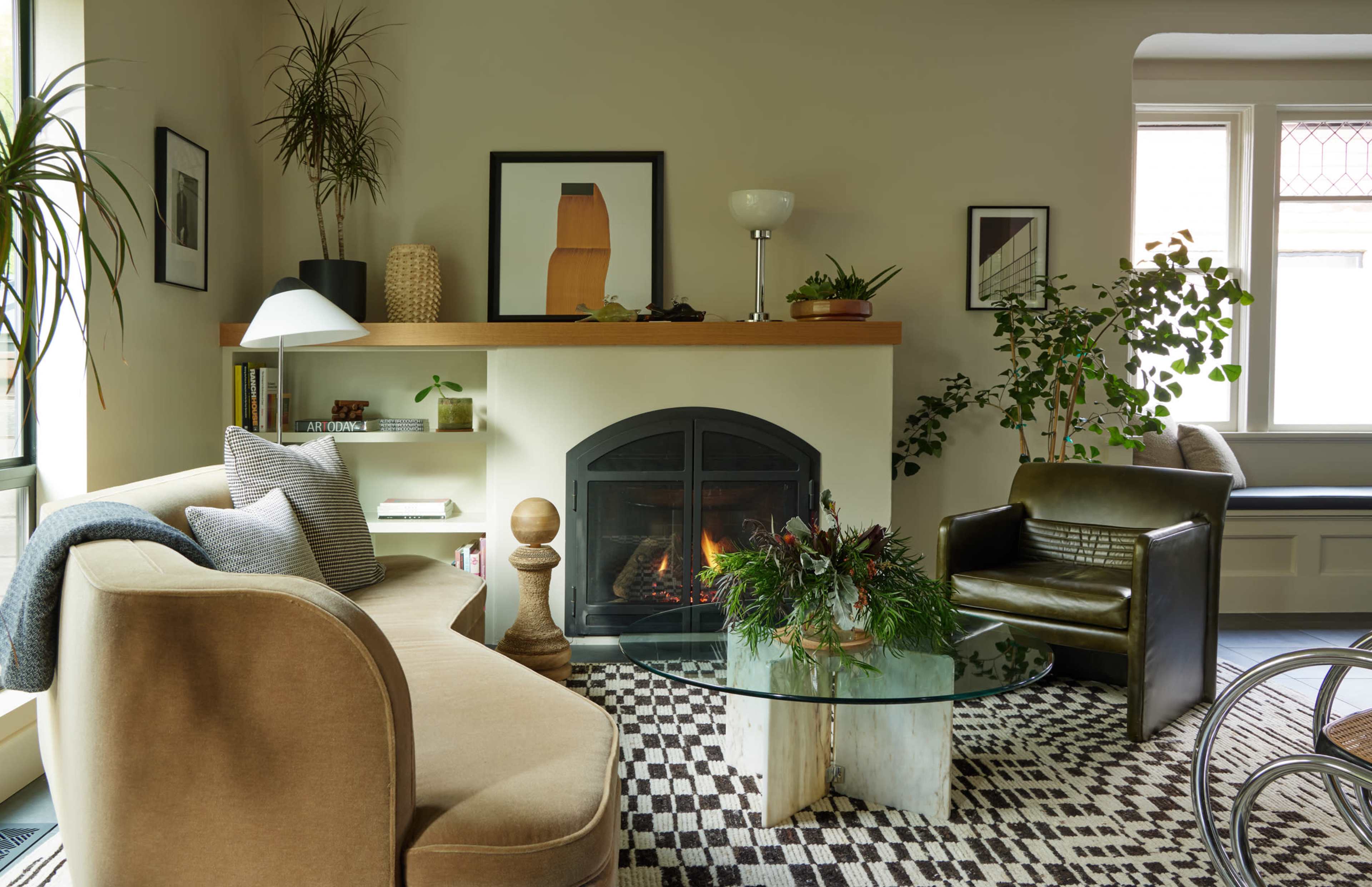 The image shows a modern living room featuring a curved beige sofa, a glass table, and a black fireplace surrounded by indoor plants.