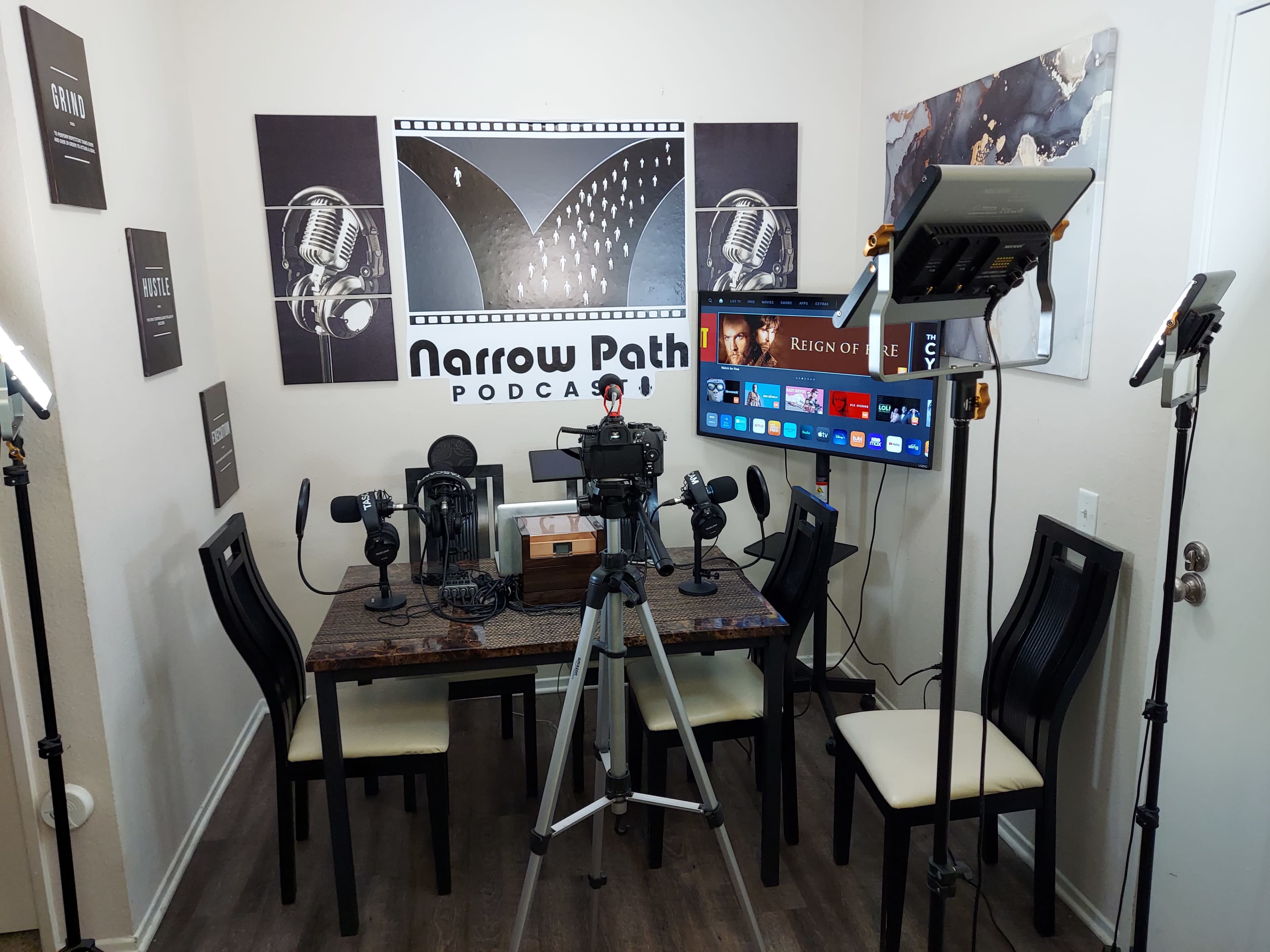 The image shows a podcast recording setup featuring a table with multiple microphones, cameras, lighting equipment, and a large display screen in a decorated room.