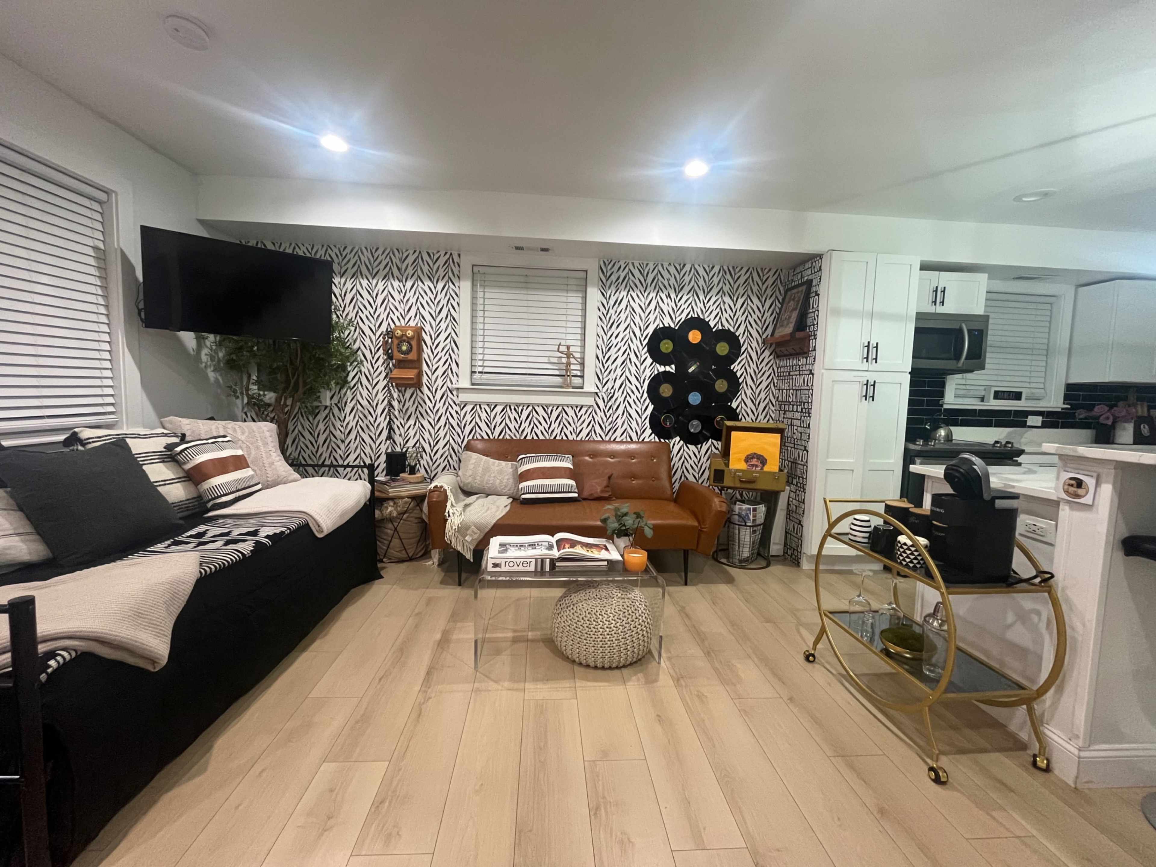 The image shows a modern living room featuring a sectional couch, a leather sofa, and a decorative wall with vinyl records and patterned wallpaper.