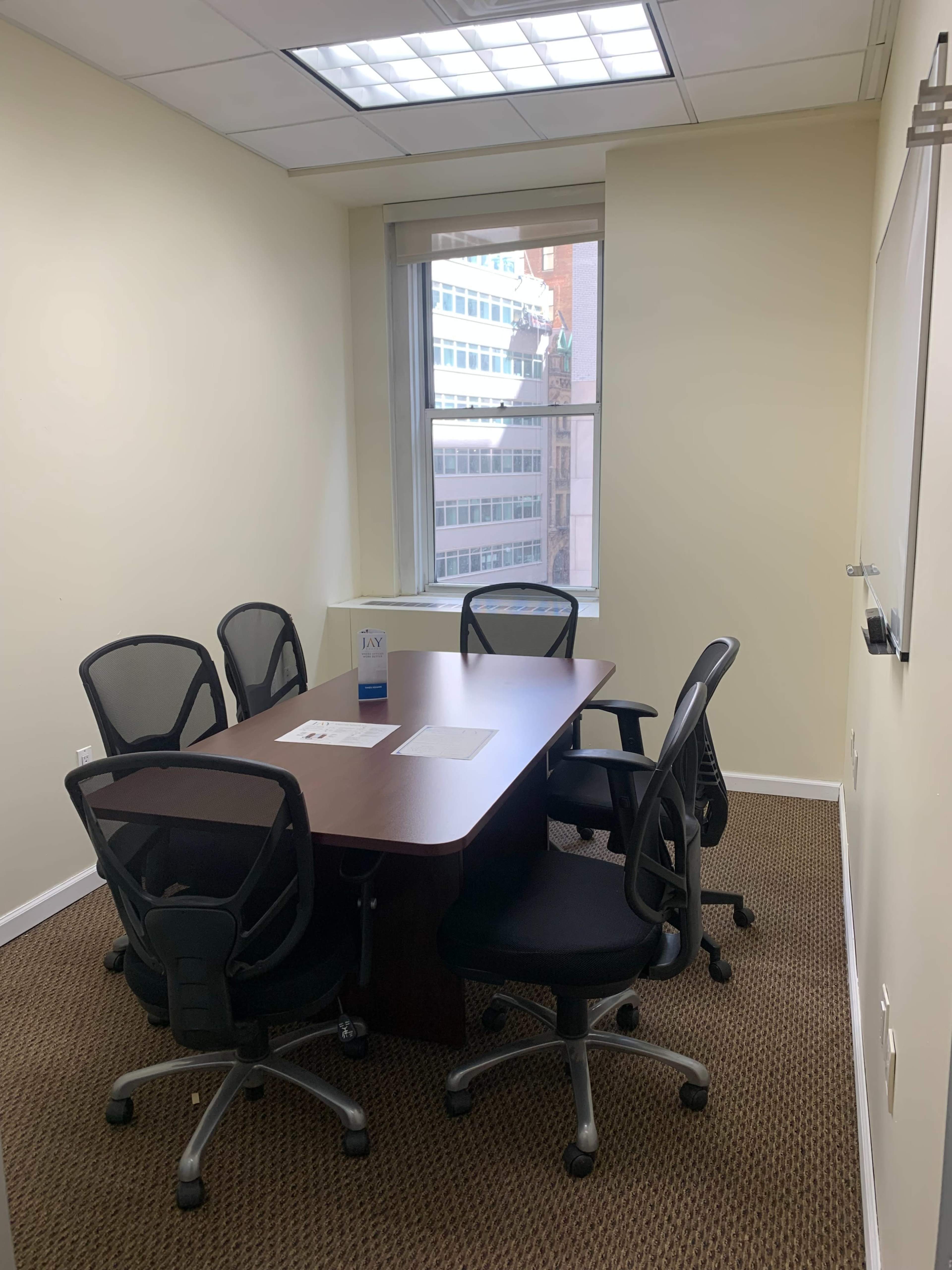 A small conference room equipped with a rectangular table and six black office chairs, featuring a window that overlooks a cityscape.