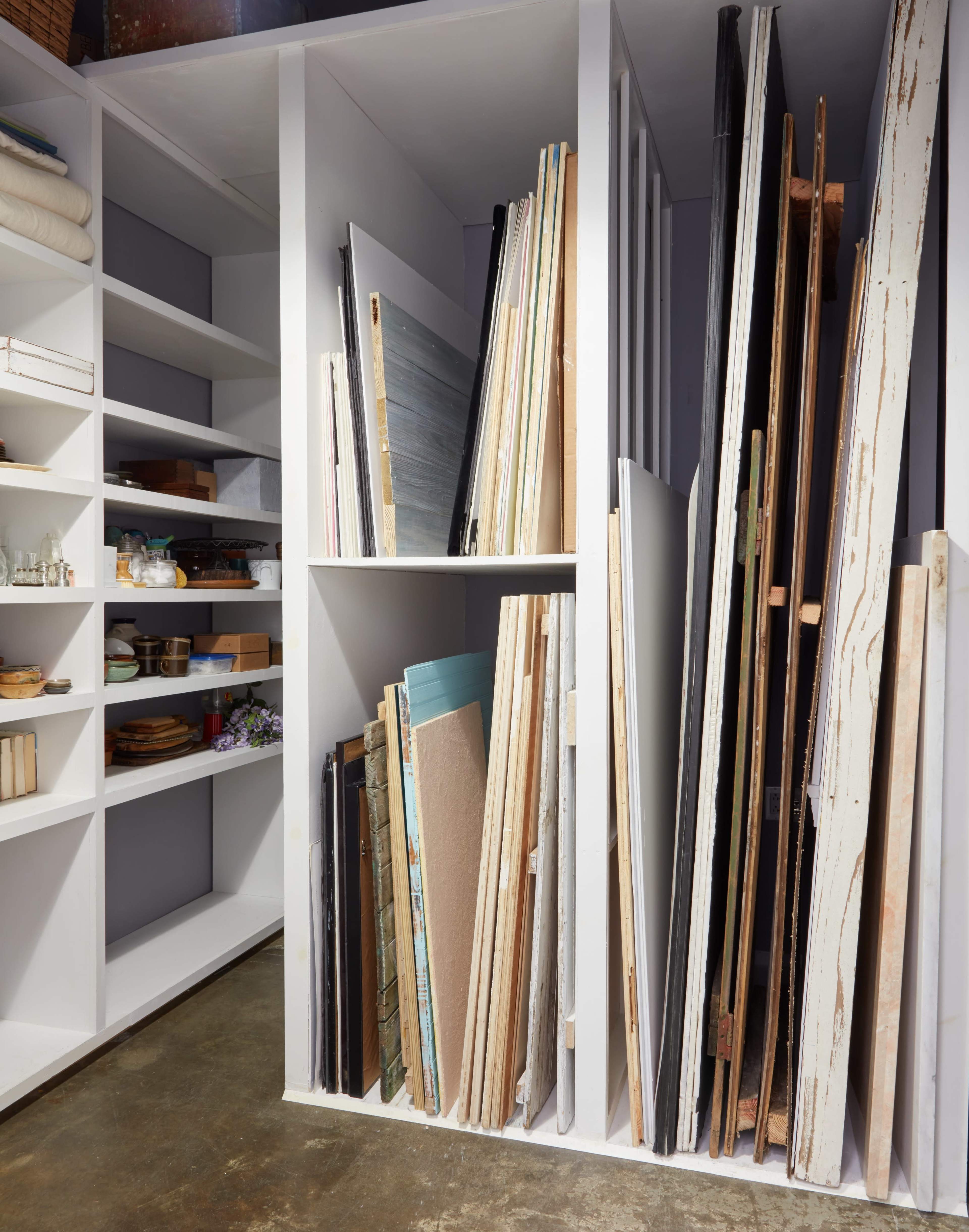 A storage area with white shelves holding various sizes of boards and canvases, alongside a collection of jars and wooden items.