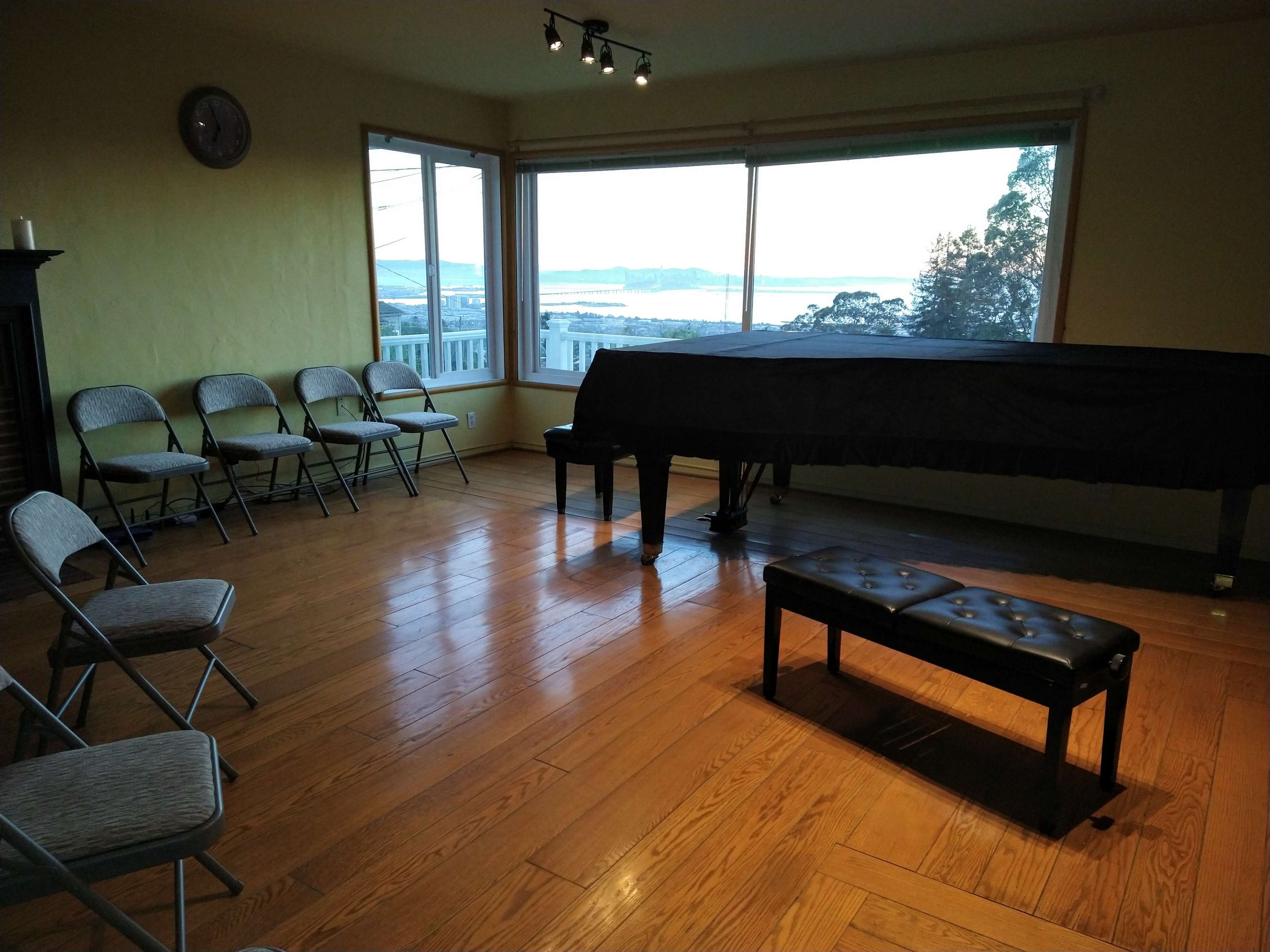 A spacious room with wooden flooring, a grand piano, a bench, and several folding chairs arranged in a semi-circle, with a large window revealing a view of the outdoors.