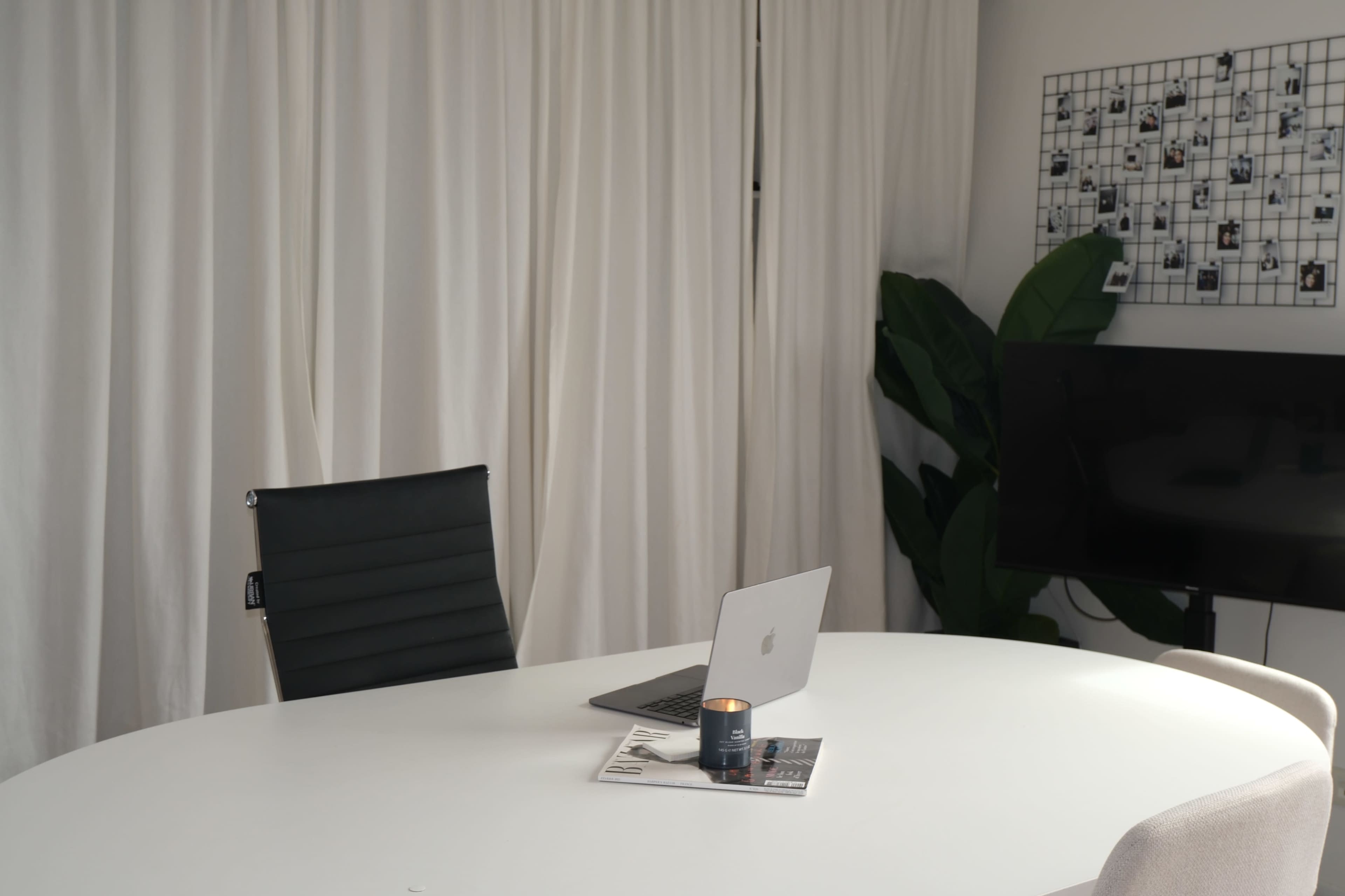 A modern conference room features a round white table, a black chair, a laptop, a candle, and green plants against a backdrop of white curtains.