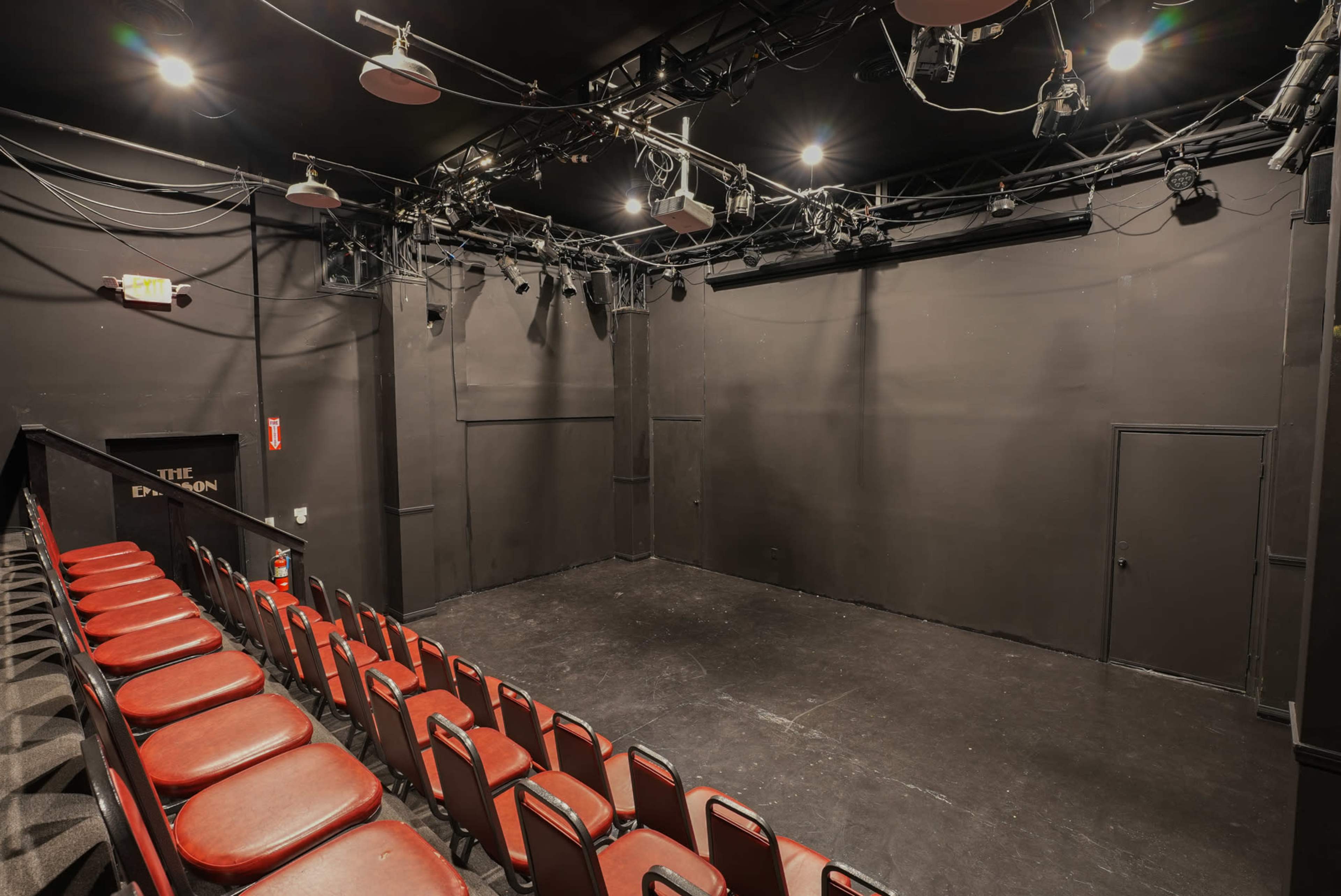 The image shows a small, black-walled theater space with an empty stage and red chairs arranged in rows.