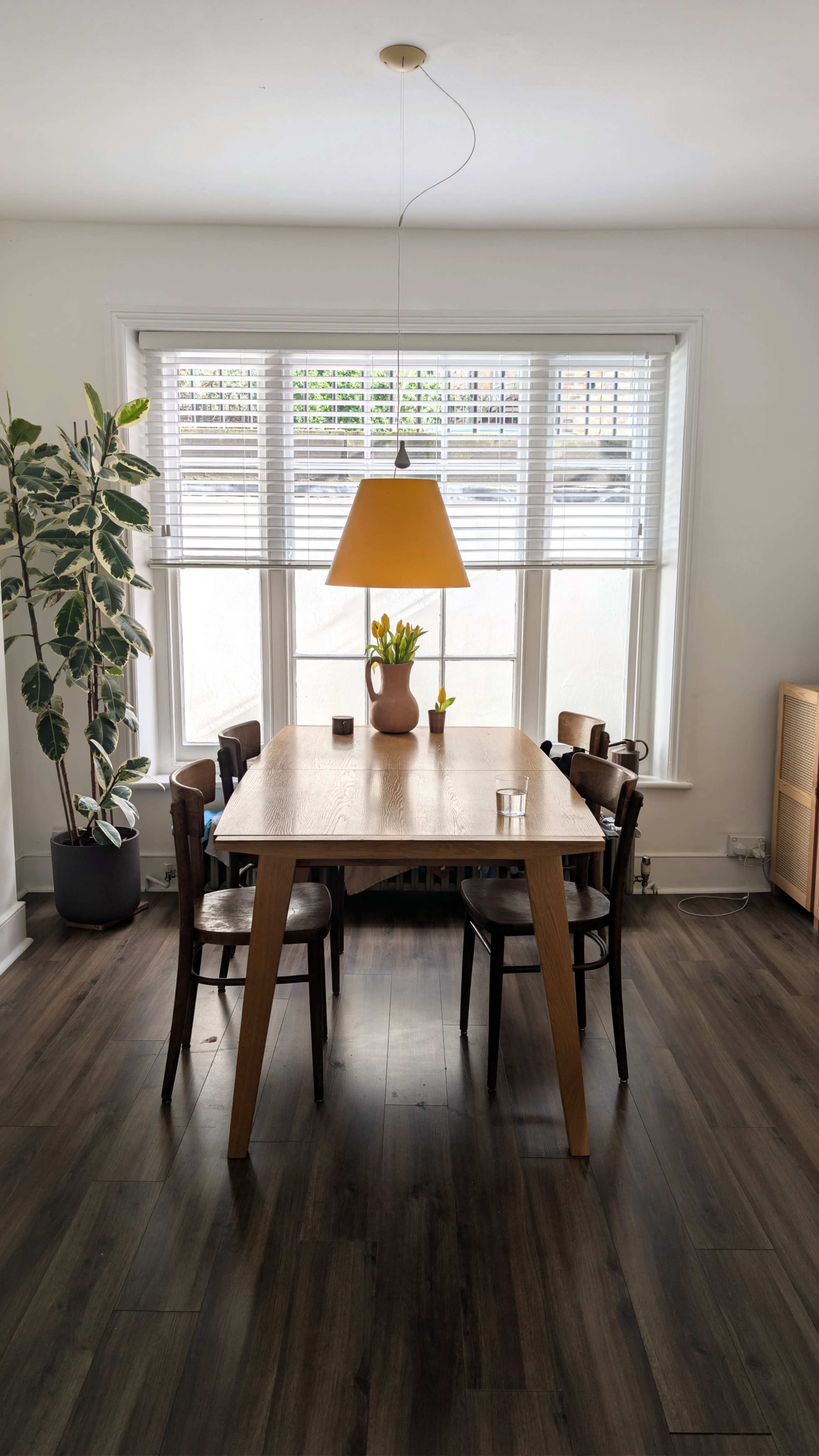 A wooden dining table surrounded by four chairs is positioned under a yellow pendant lamp in a well-lit room with large windows and a potted plant.